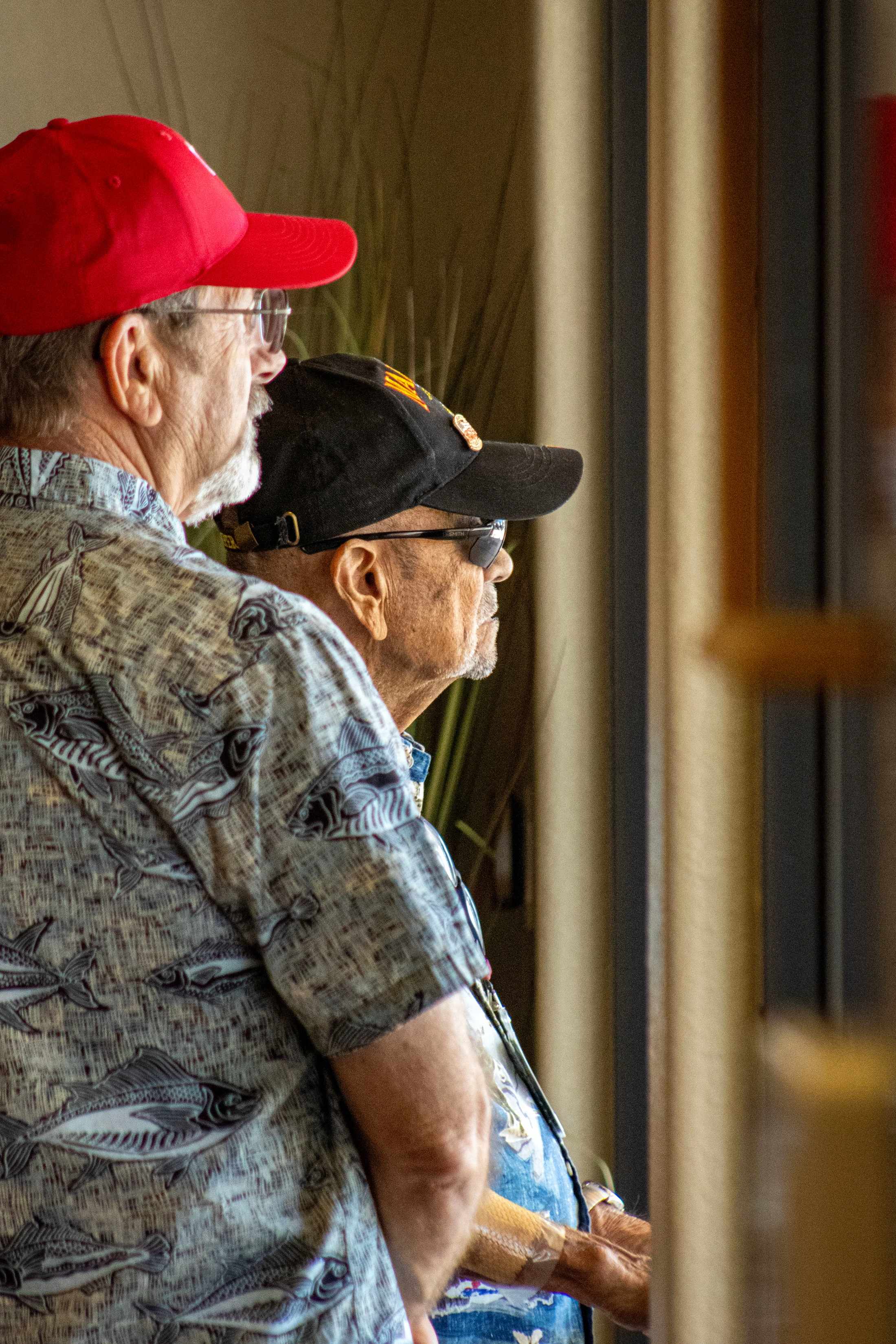 A profile view of two older men standing together and looking to the right. The man in front wears a red cap and a fish-patterned shirt.
