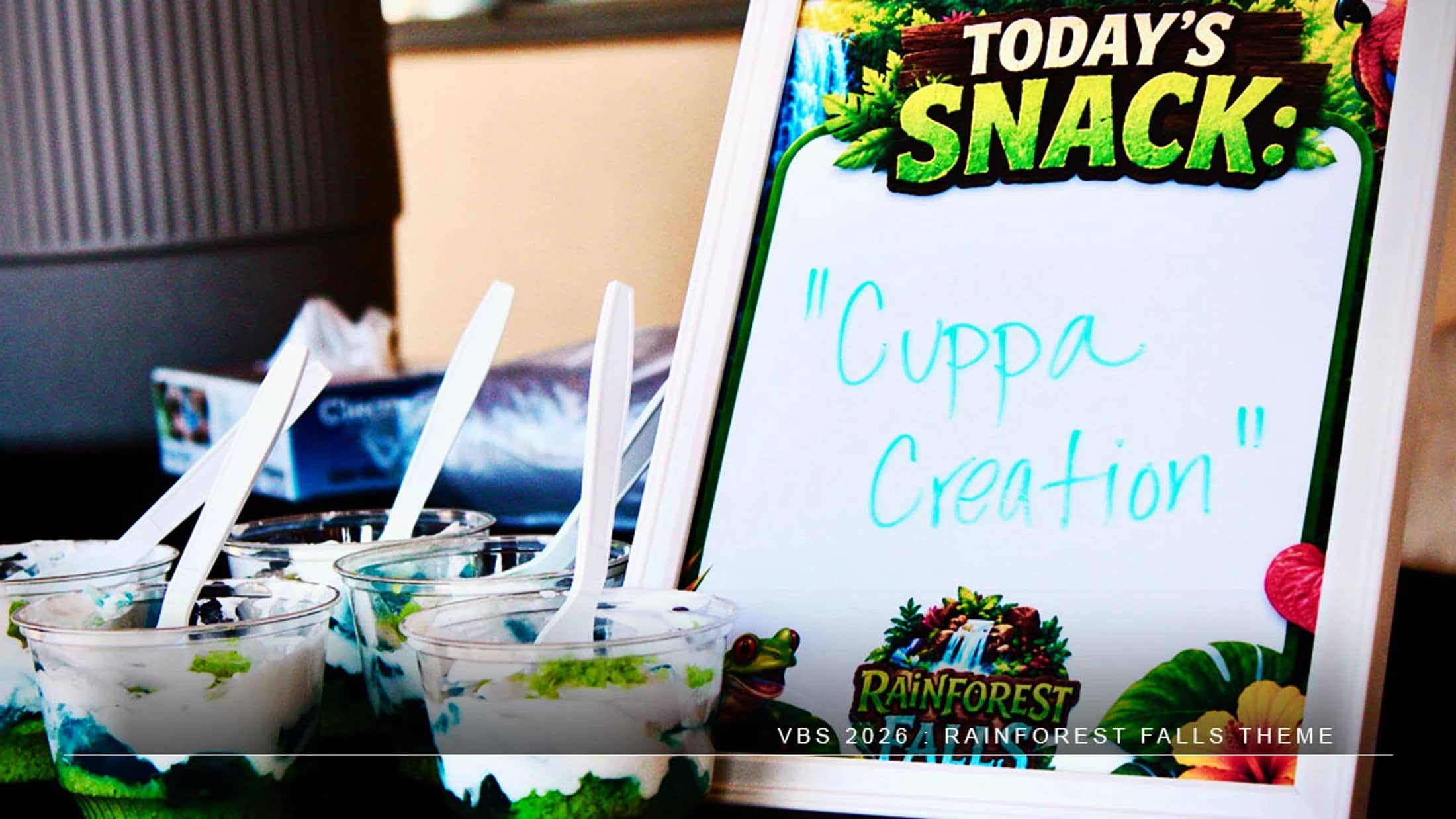 A whiteboard sign reads 'Today's Snack: Cuppa Creation' next to layered dessert cups with spoons for a rainforest-themed Vacation Bible School event.