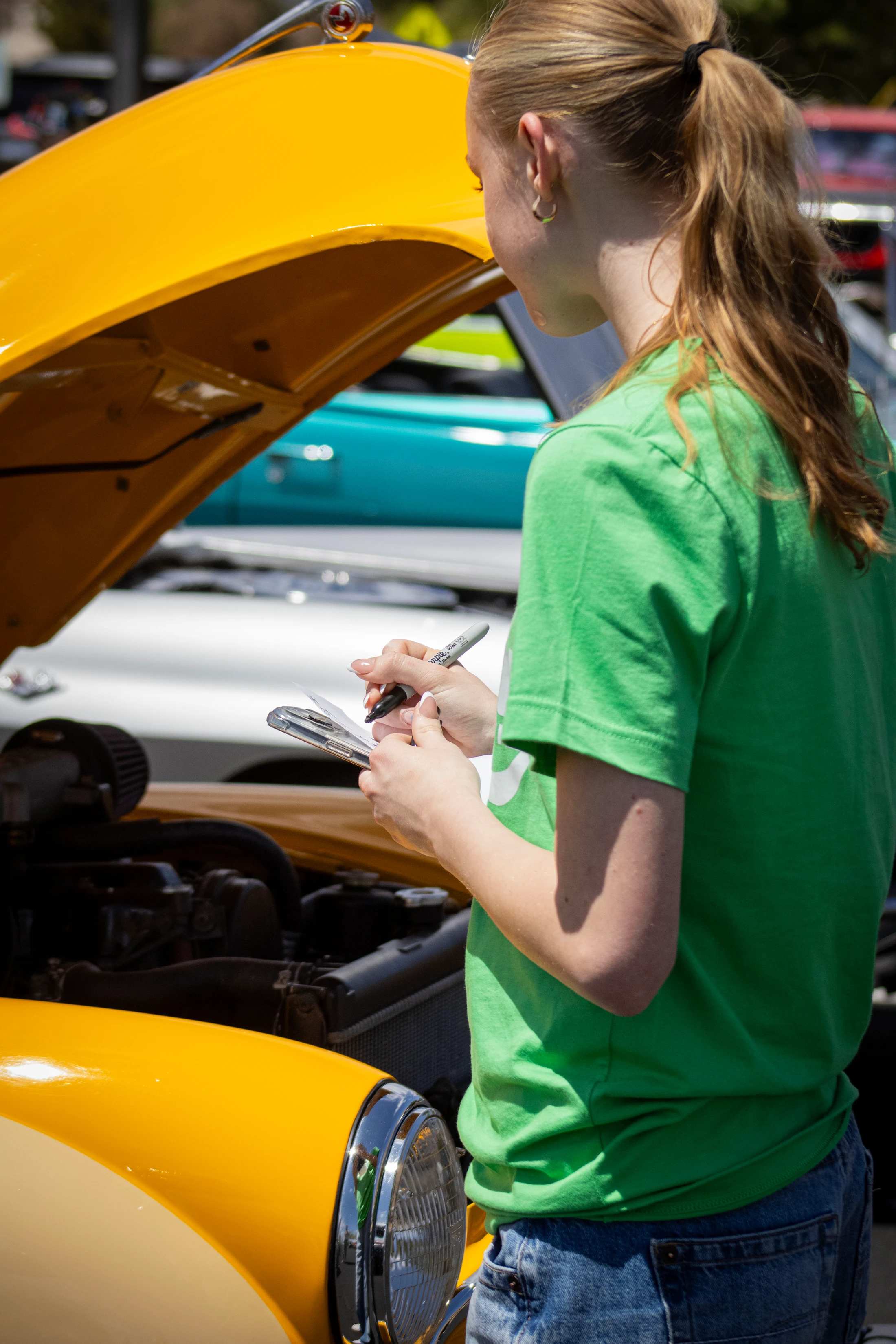 A young woman with a ponytail, wearing a green shirt, writes on a clipboard while inspecting the engine of a vintage yellow car with the hood open.