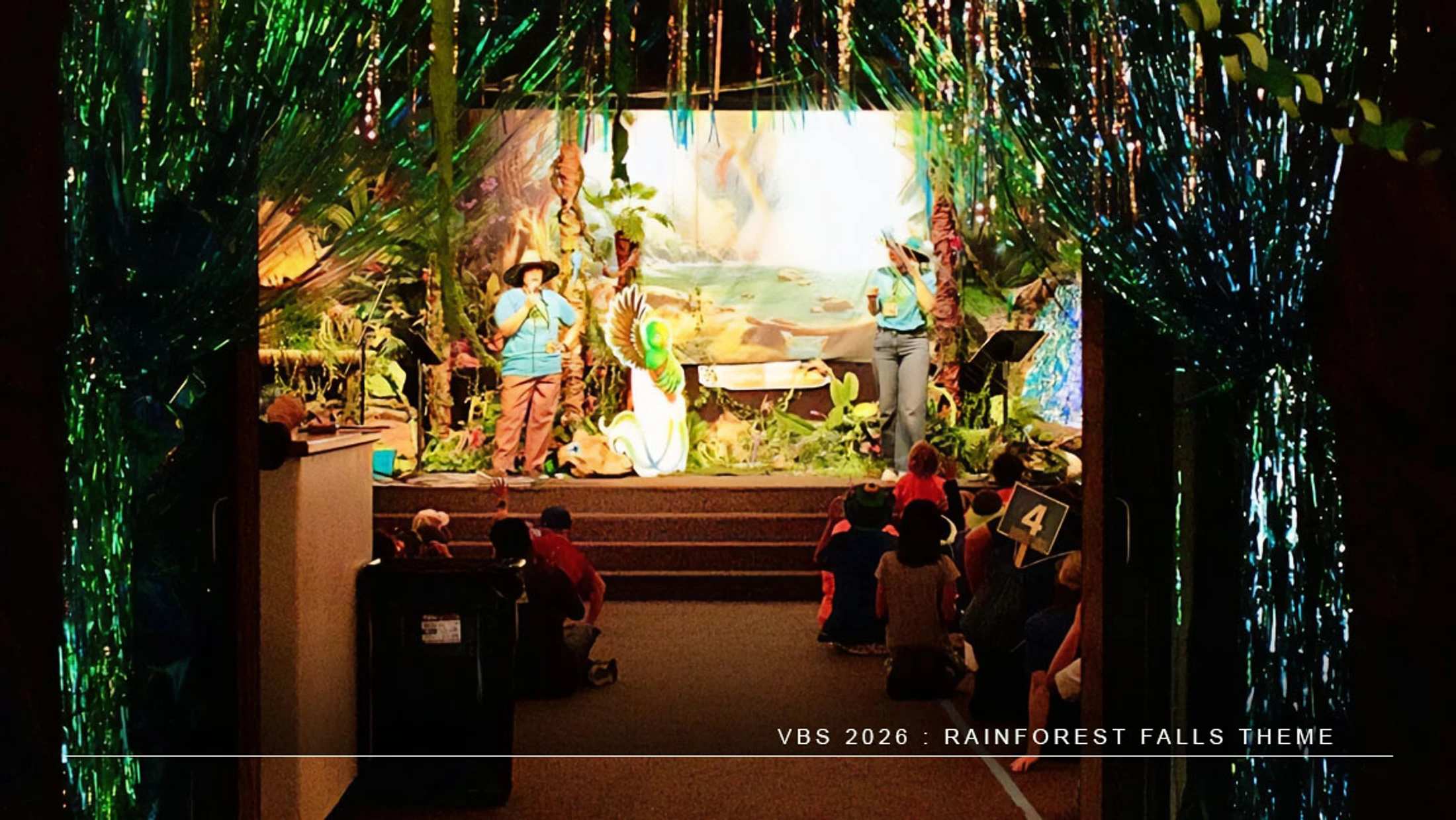 Children sit on the floor watching two adults on a stage decorated with a rainforest theme, framed by green tinsel curtains, for a VBS event.