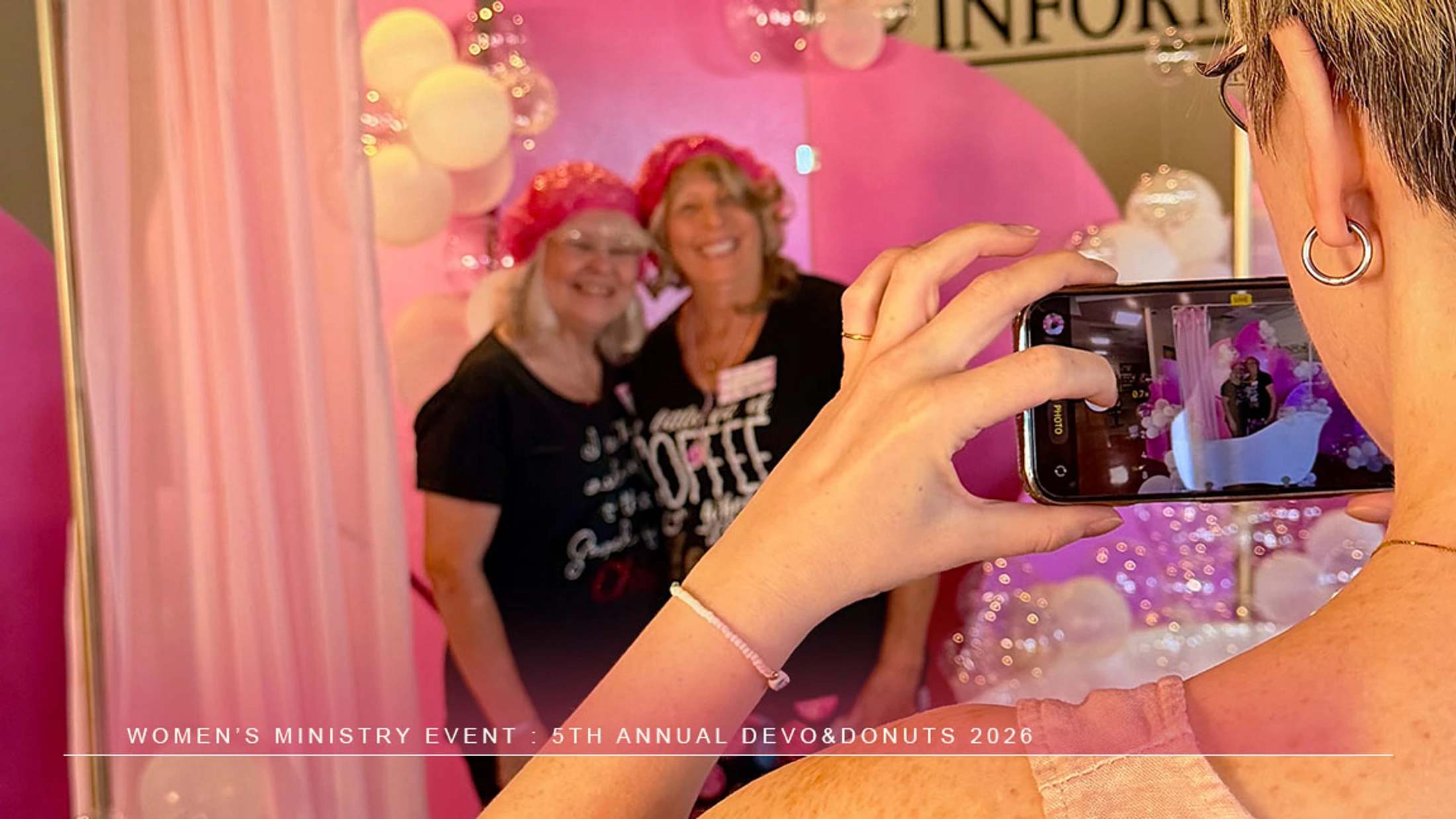 An over-the-shoulder shot of a woman taking a smartphone photo of two smiling women in pink hats posing in front of a pink balloon backdrop.