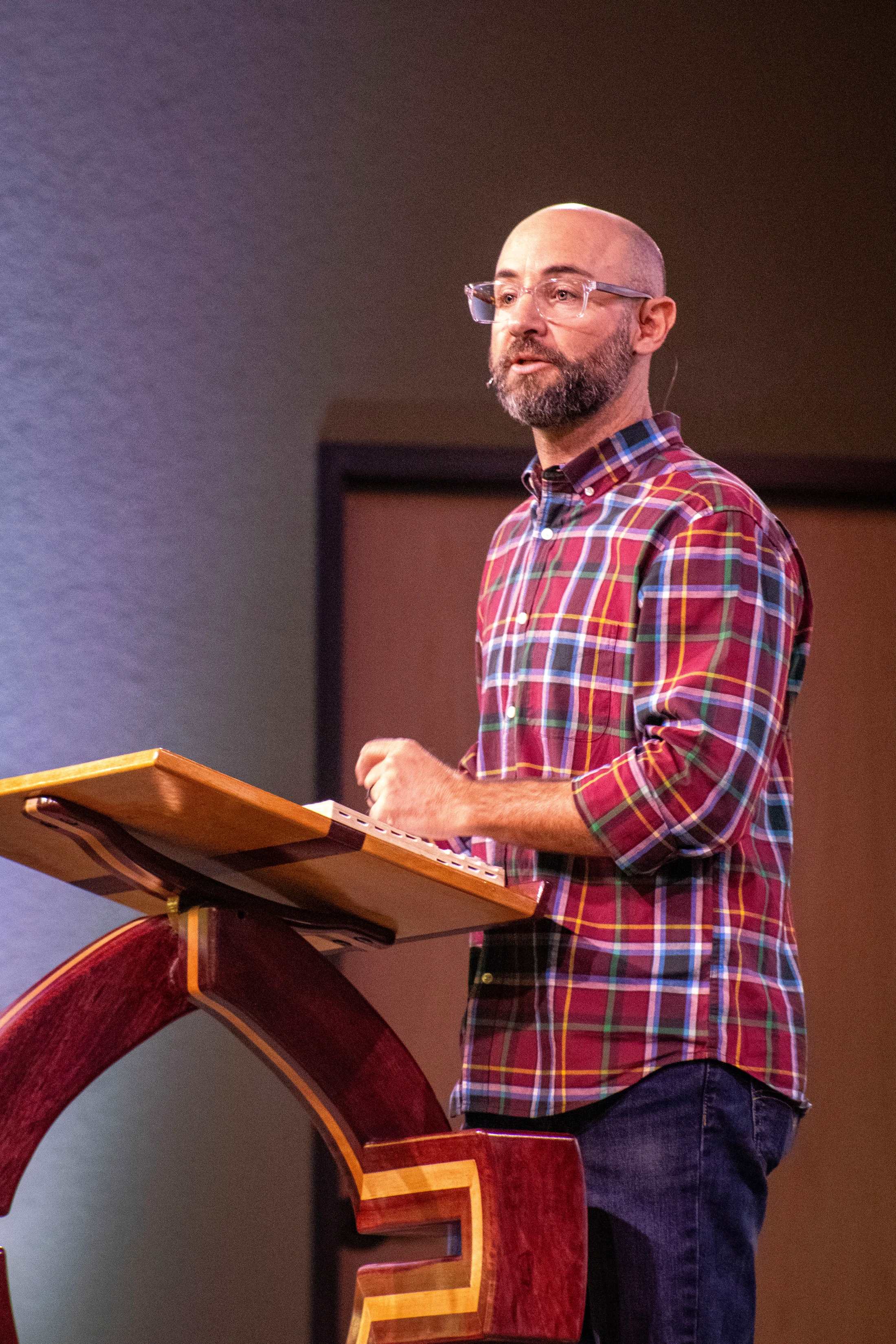 A bald man with a beard and glasses, wearing a colorful plaid shirt, stands behind a wooden lectern while speaking. His hands are on an open book.
