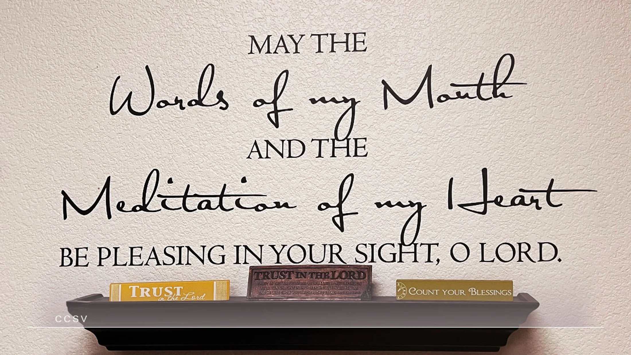 A textured wall with a Christian prayer written in black script. Below, a shelf holds signs reading 'Trust in the Lord' and 'Count Your Blessings'.