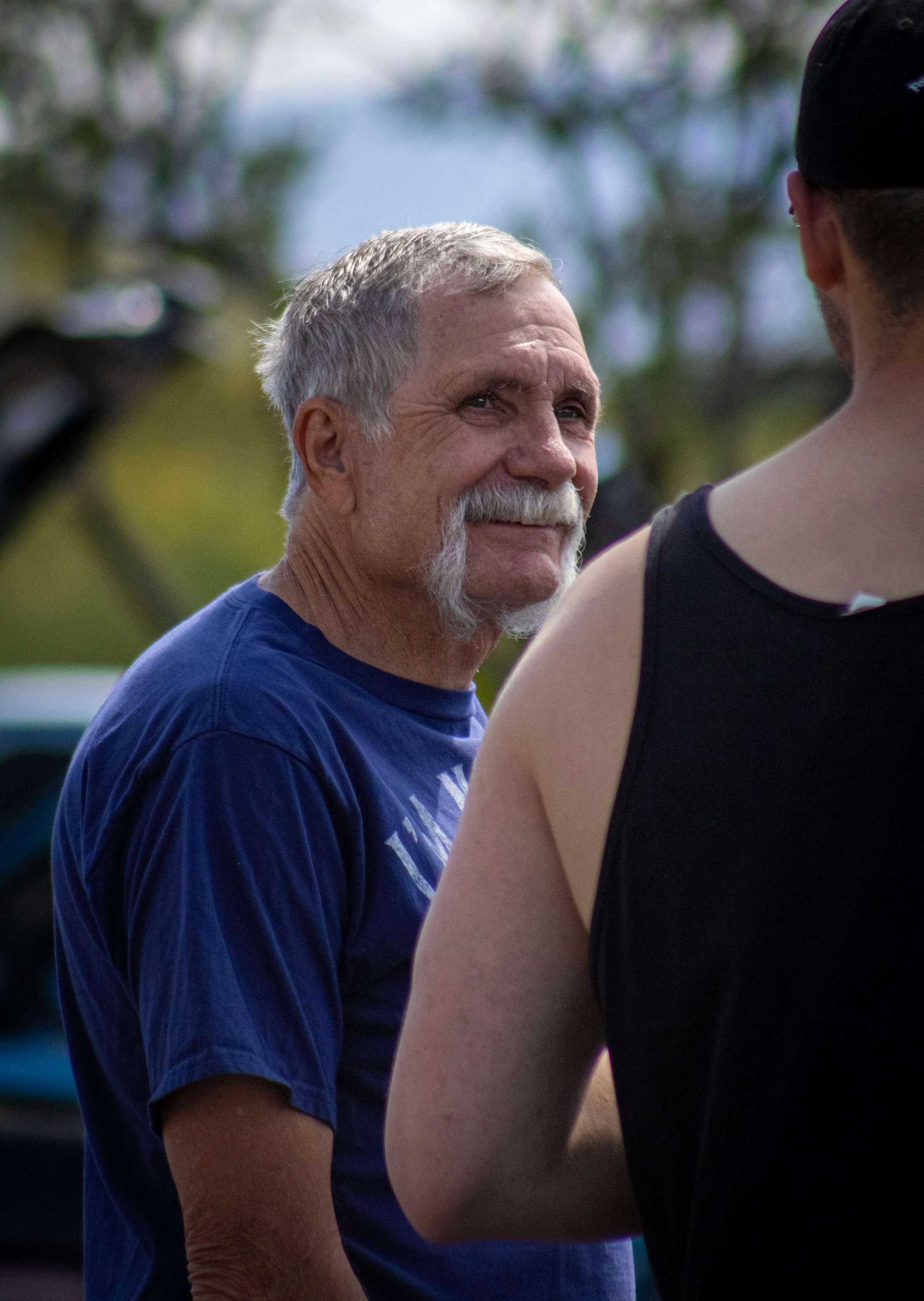 An older white man with grey hair and a large handlebar mustache smiles while talking to another person. He wears a blue t-shirt and is seen from the chest up.