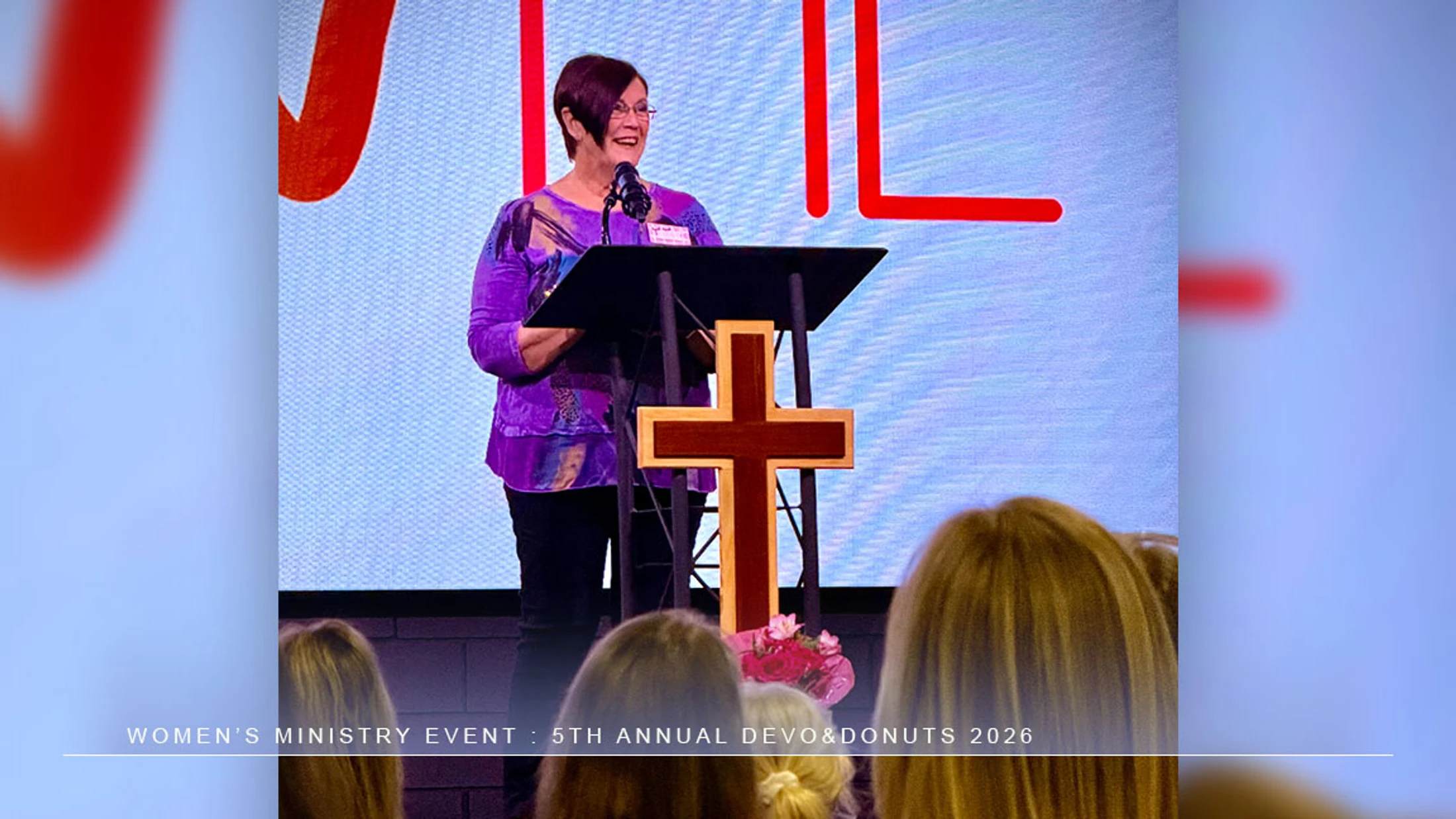 A woman with short dark hair and a purple shirt smiles as she speaks at a podium with a wooden cross on the front, addressing an audience at a church event.