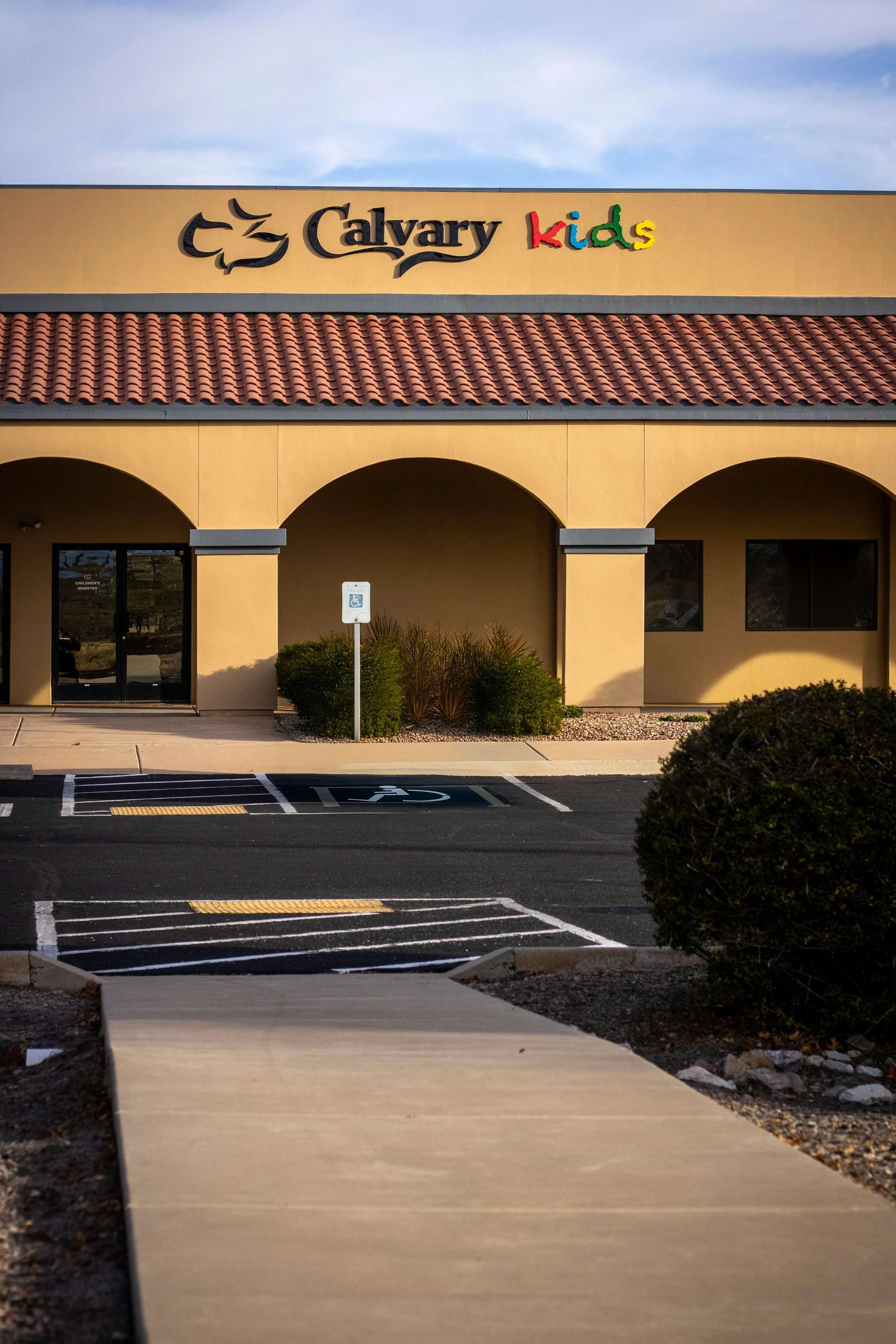 The front of a yellow stucco building with a red tile roof and arched entryways. A colorful sign reads 'Calvary Kids'. A parking lot is in the foreground.