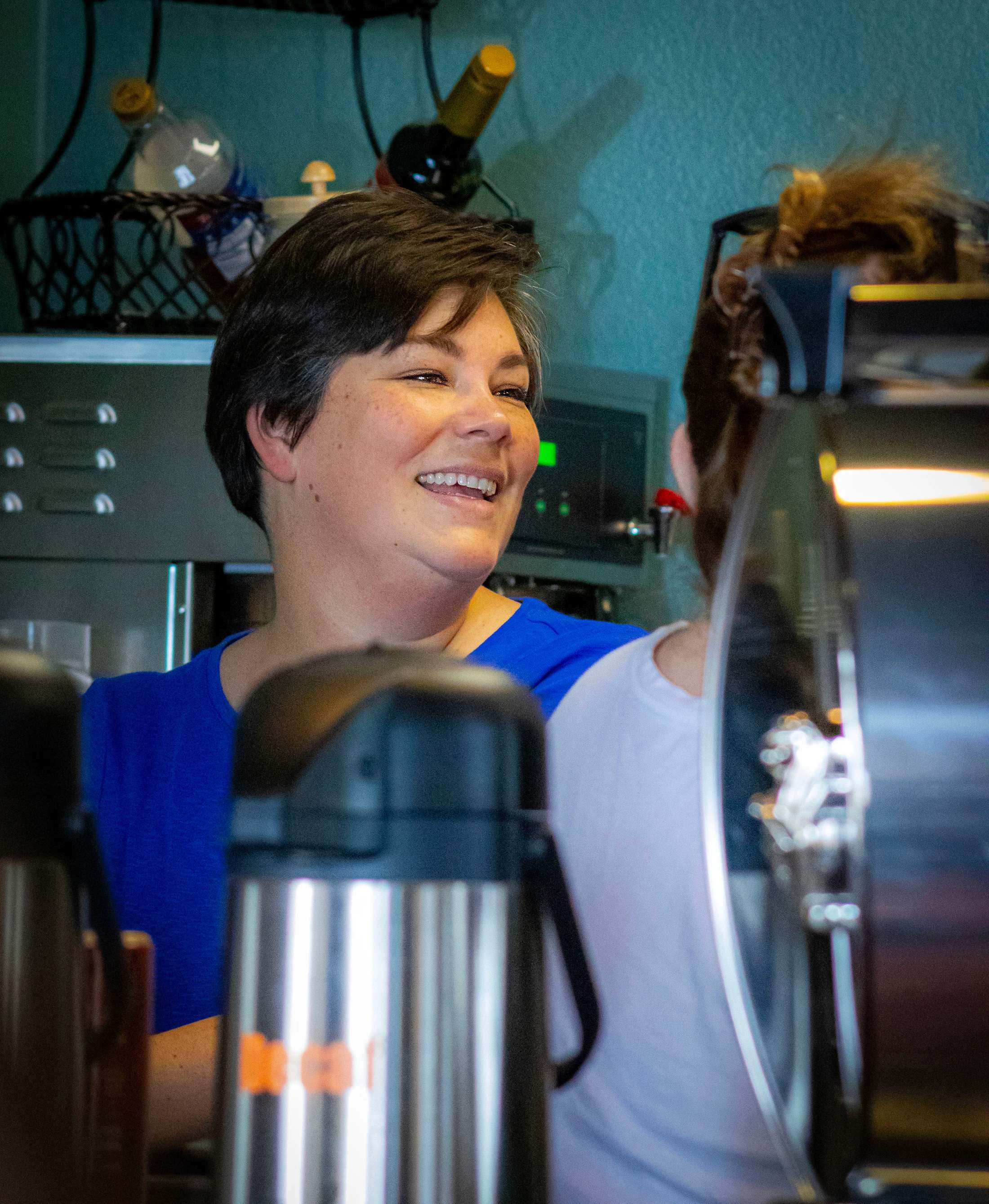 A woman with short dark hair and a blue shirt smiles as she talks to another person in a cafe, with metal coffee carafes in the blurry foreground.