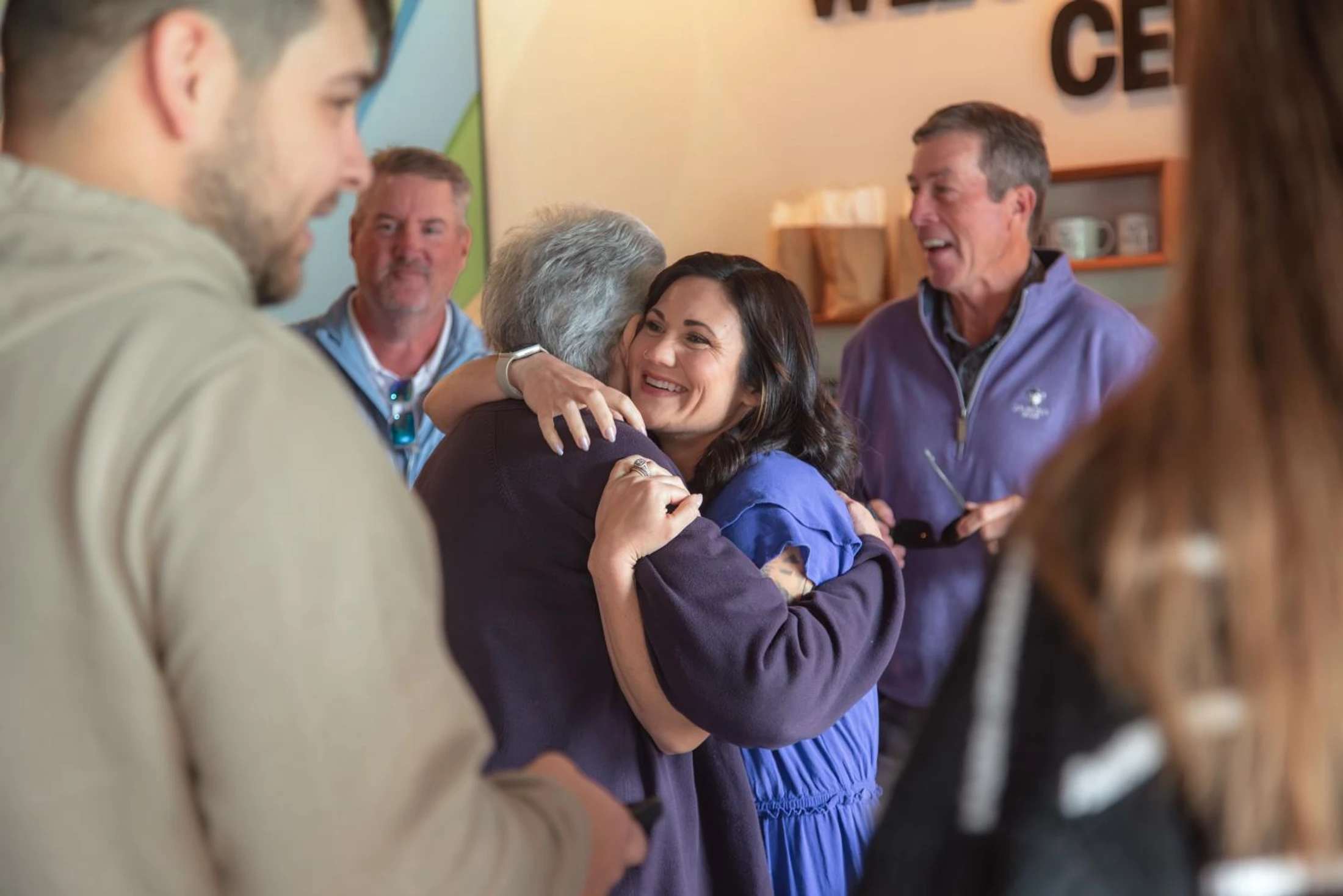 A smiling woman in a blue dress hugs an older person. They are surrounded by other people at what appears to be a happy indoor get-together.