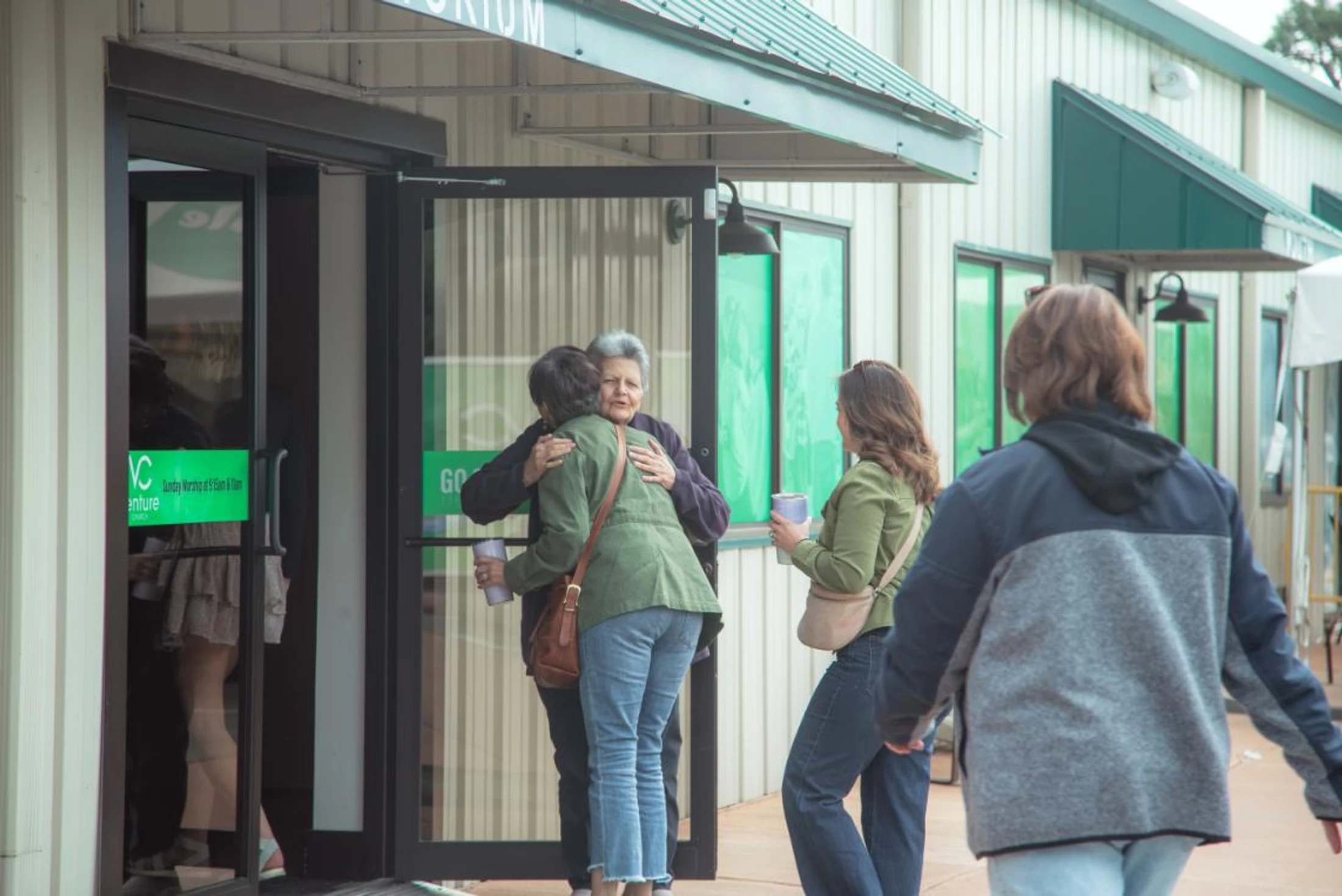 Two older women share a warm hug at the entrance to a church auditorium, while other people stand nearby.