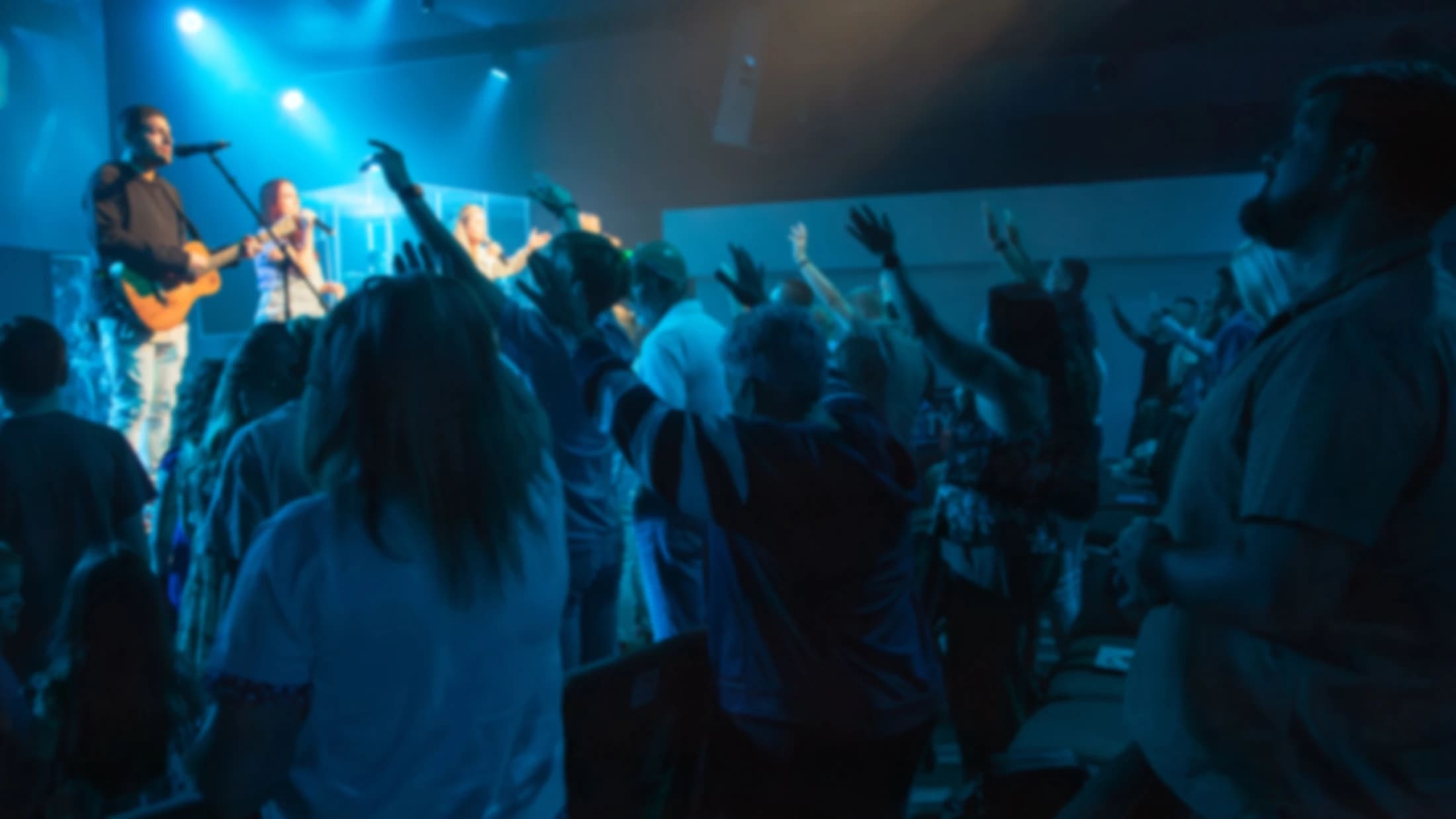 A blurry, low-light photo of a congregation with their hands raised in worship. A band performs on stage under blue lights.