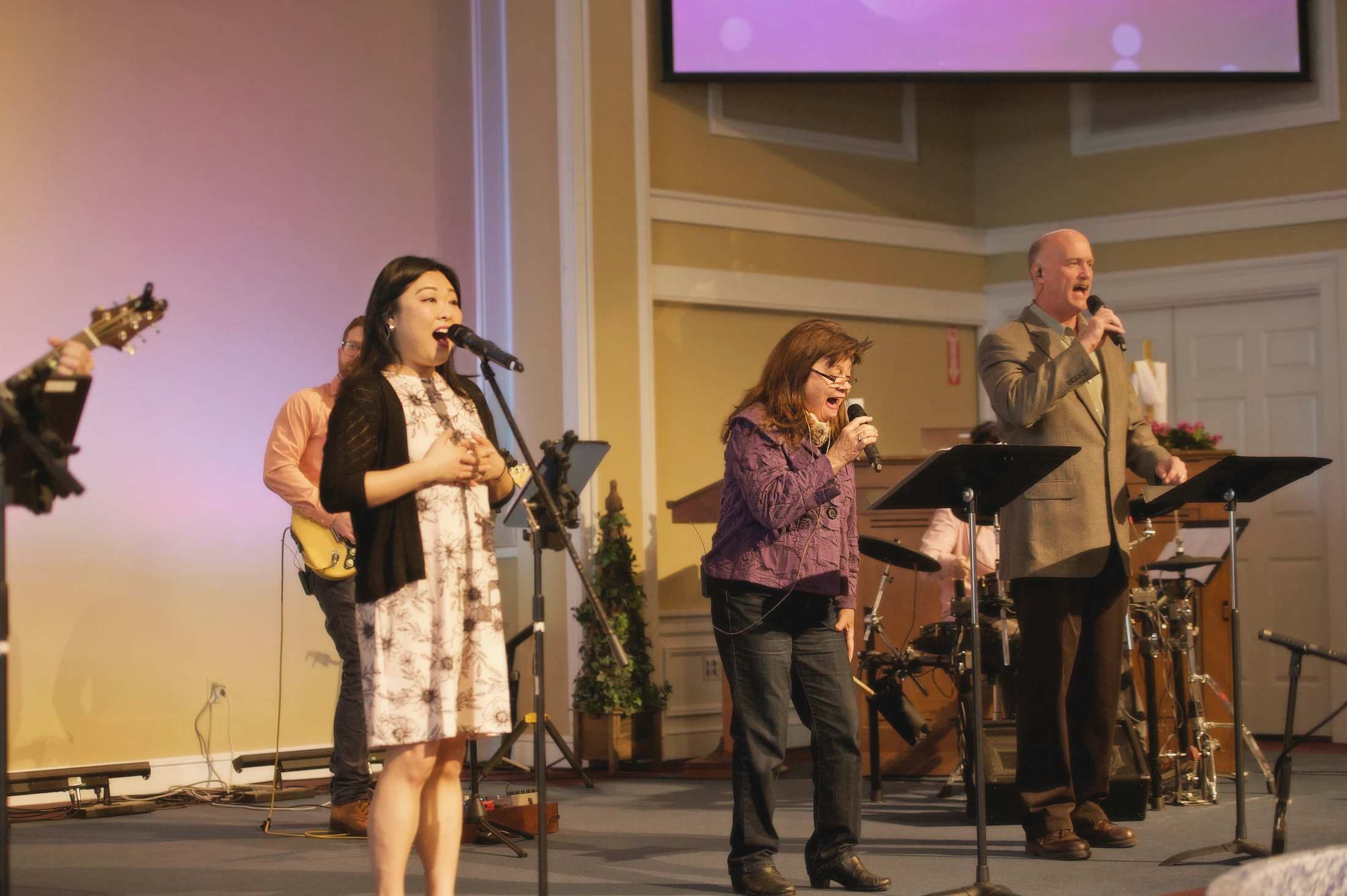A band performs on stage with three lead singers, two women and a man, singing into microphones. Other band members are visible in the background.