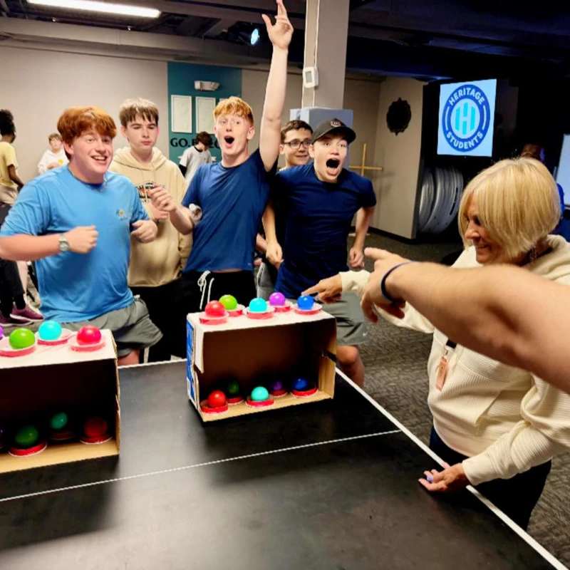 A group of teenage boys with excited expressions cheer while playing a game involving pressing colorful buttons, with a woman smiling and pointing.
