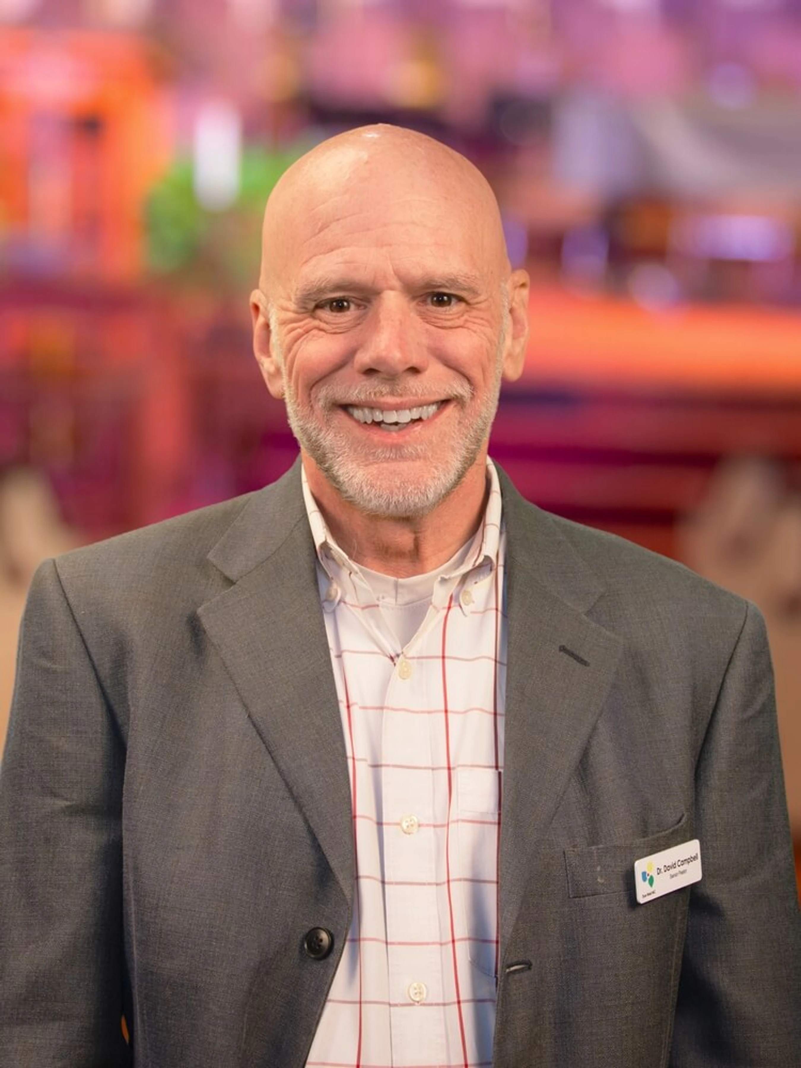 A head and shoulders portrait of a smiling, bald man with a gray beard. He is wearing a gray blazer over a plaid shirt and has a name tag on his lapel.