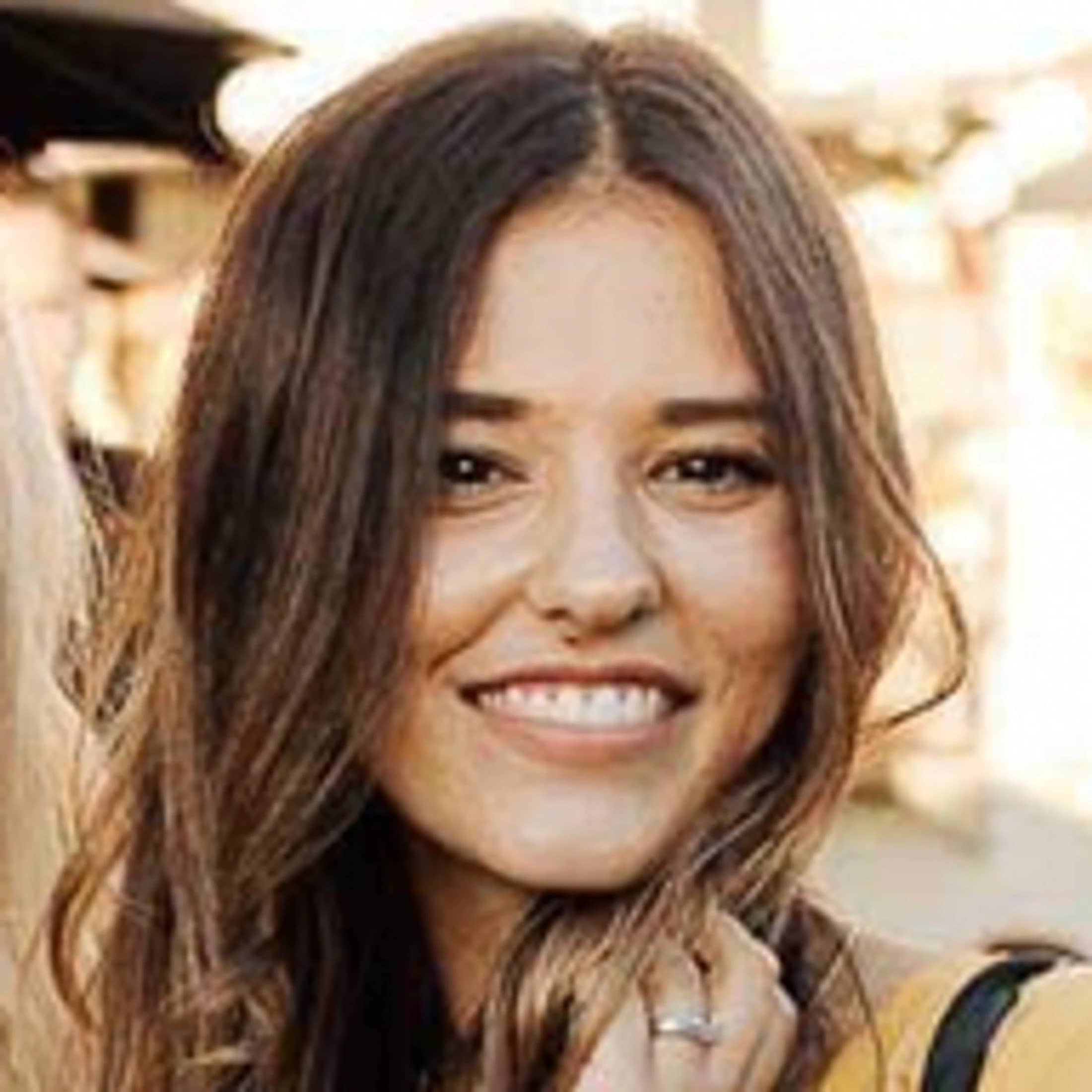A close-up headshot of a woman with long, wavy brown hair and brown eyes. She is smiling broadly, looking directly at the camera.