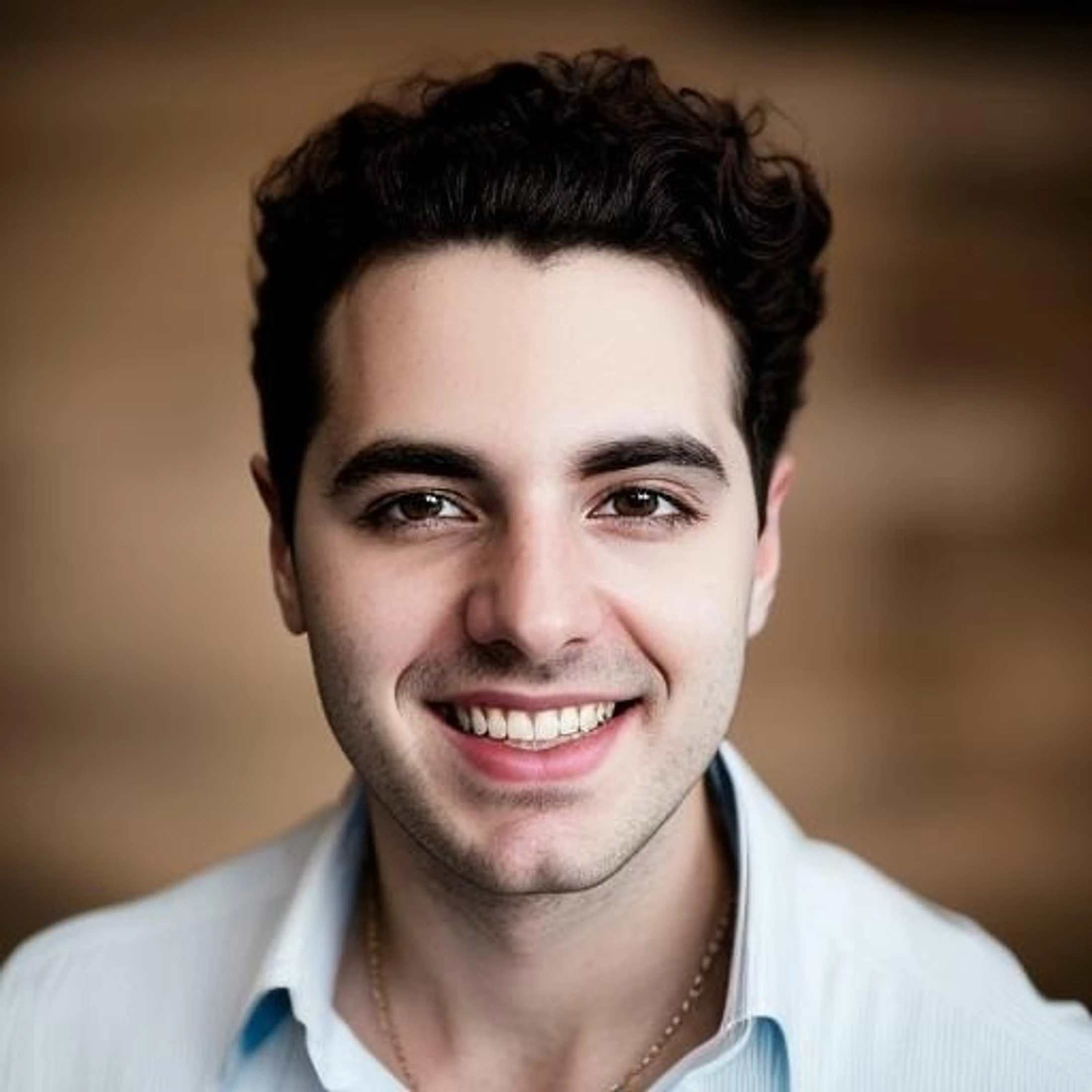 A headshot of a young man with dark wavy hair and brown eyes, smiling warmly at the camera. He is wearing a light blue collared shirt and a gold chain.