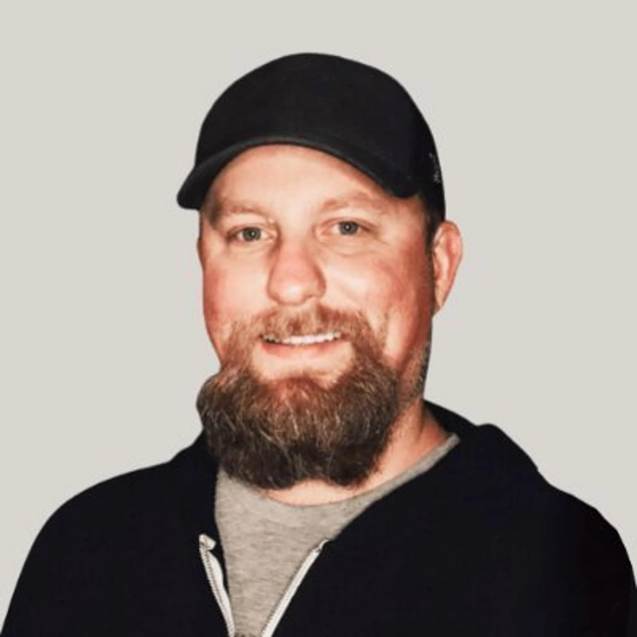 A friendly-looking white man with a full brown beard and blue eyes smiles at the camera. He wears a black baseball cap and a black zip-up hoodie.