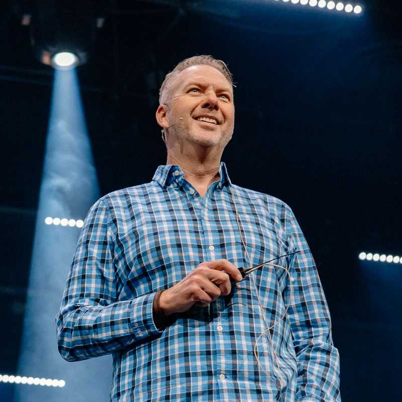 A smiling middle-aged man in a blue and black plaid shirt stands on a stage. A spotlight shines behind him as he holds part of his audio equipment.