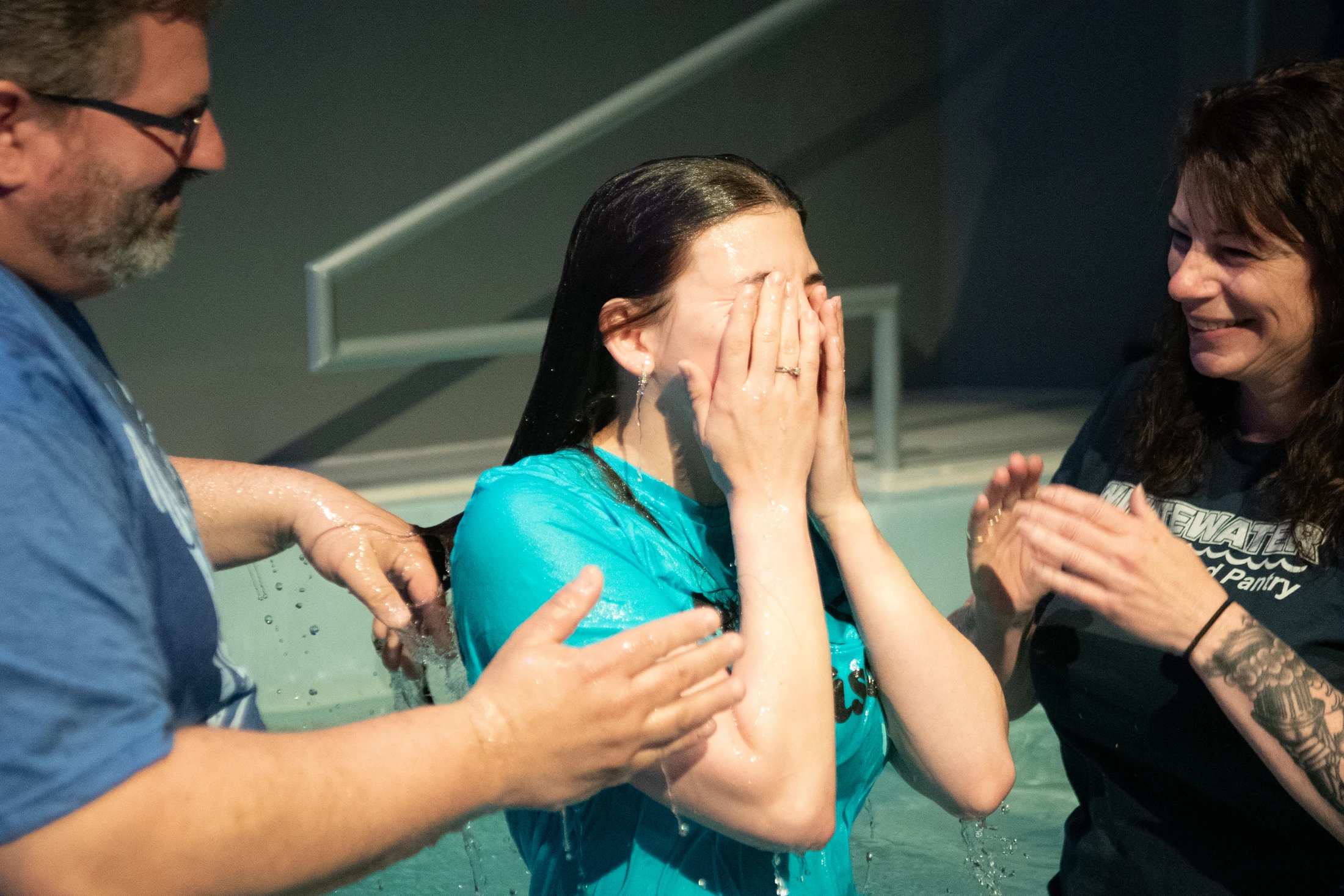 A young woman in a turquoise shirt covers her face with her hands as she comes up from the water in a baptismal pool, assisted by a man and another woman.