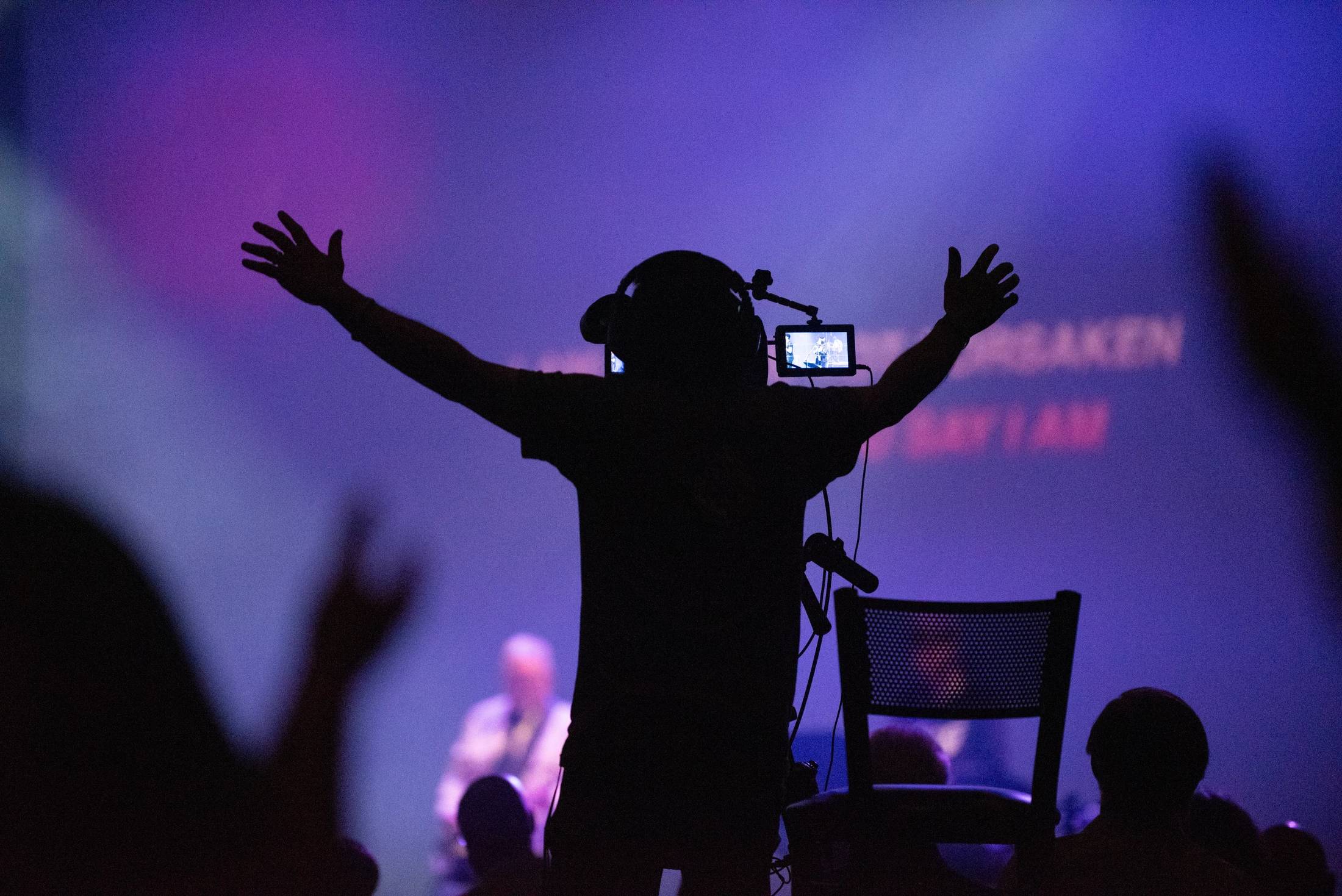 A silhouette of a camera operator wearing headphones, with his arms raised in worship in front of a purple-lit stage during a live event.