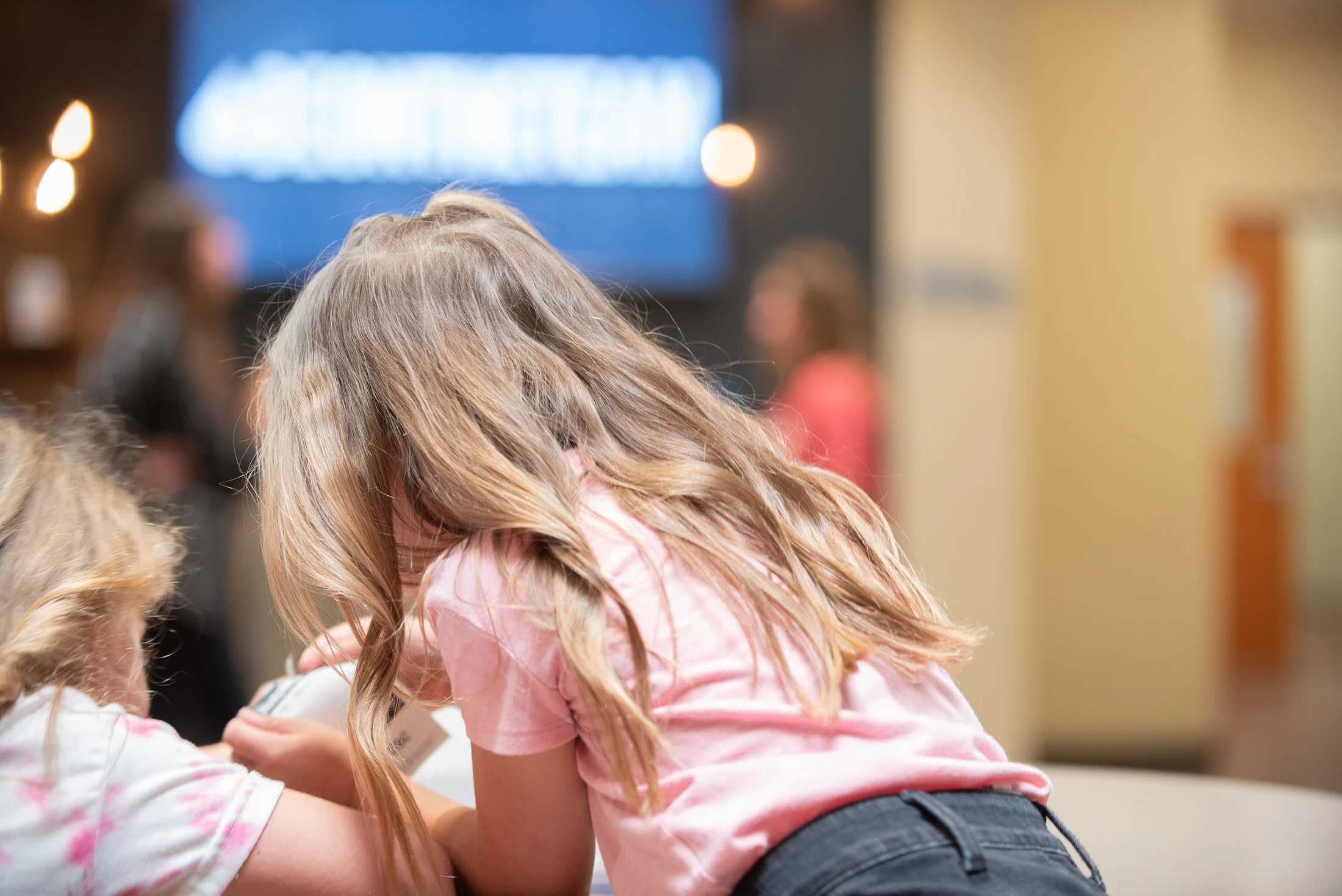 The back of a young girl with long, wavy blonde hair wearing a pink t-shirt. She is leaning forward indoors, with the background softly blurred.