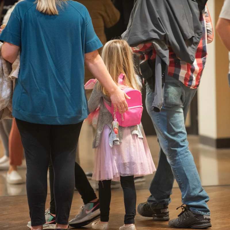 From behind, a young girl with a pink backpack walks between a woman and a man. The woman's hand rests gently on the girl's back as they walk together.