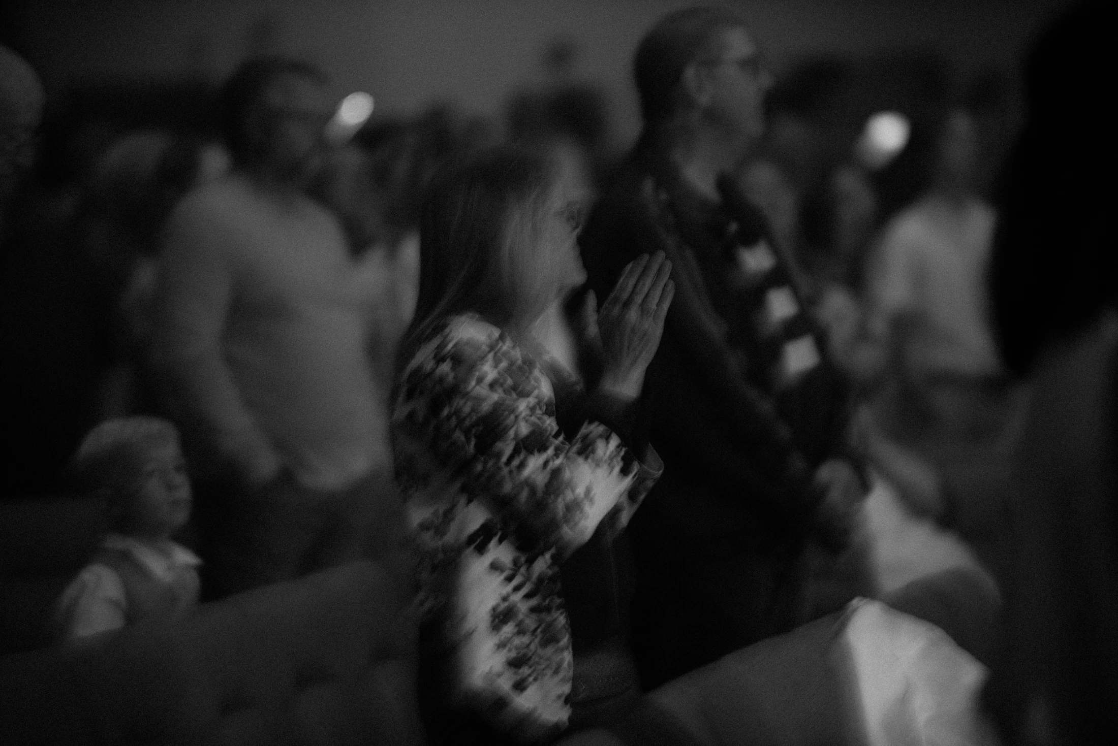 A grainy, black-and-white, blurry photo of a woman in profile with her hands clasped. She is surrounded by other out-of-focus people in a crowd.