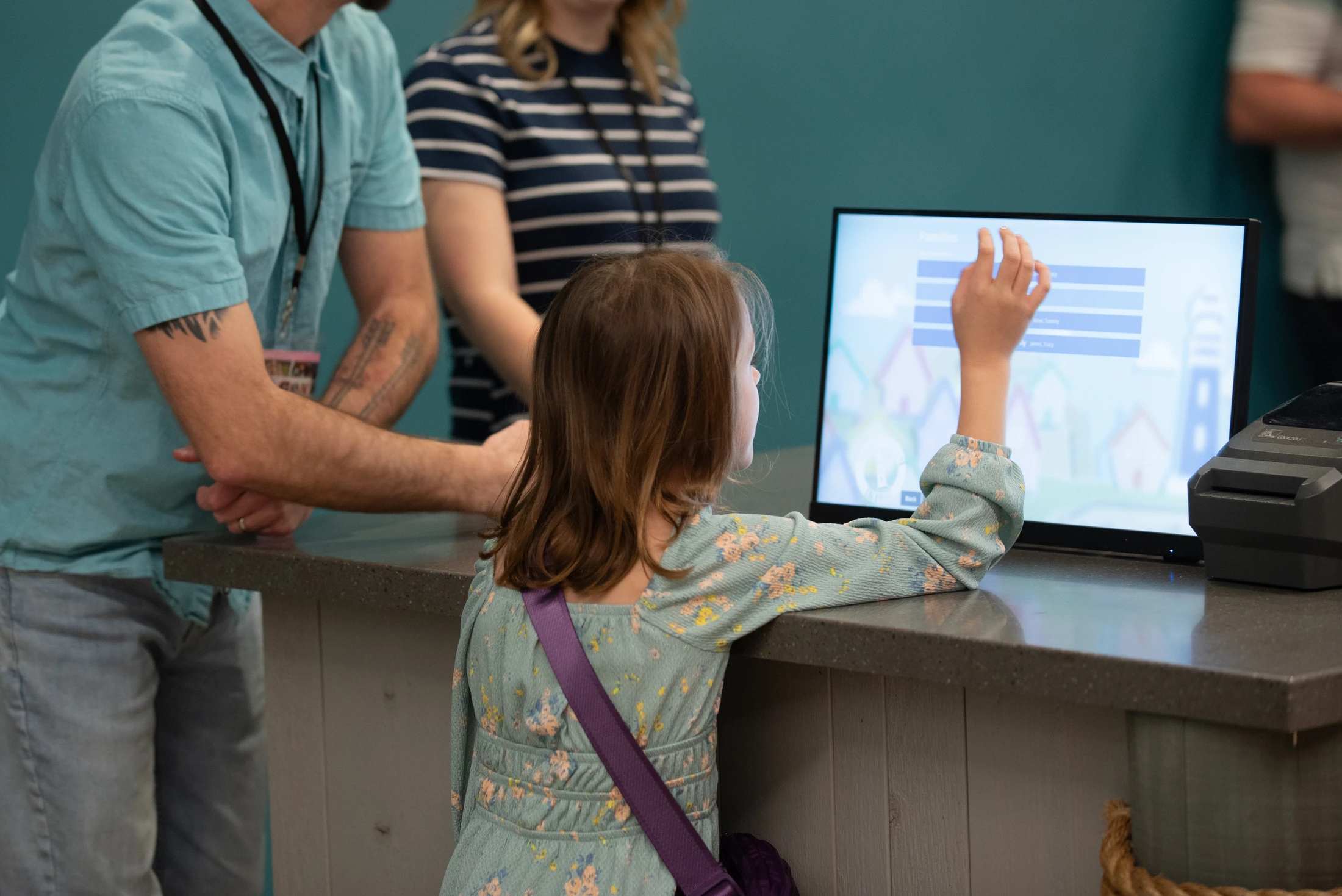 A young girl with her parents behind her uses her finger to interact with a touchscreen monitor at a check-in counter for an event or service.