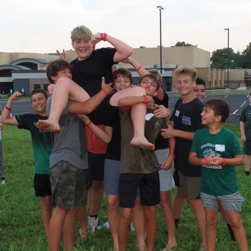 A group of teenage boys smile and pose on grass, with several lifting one boy onto their shoulders who is flexing and grinning.