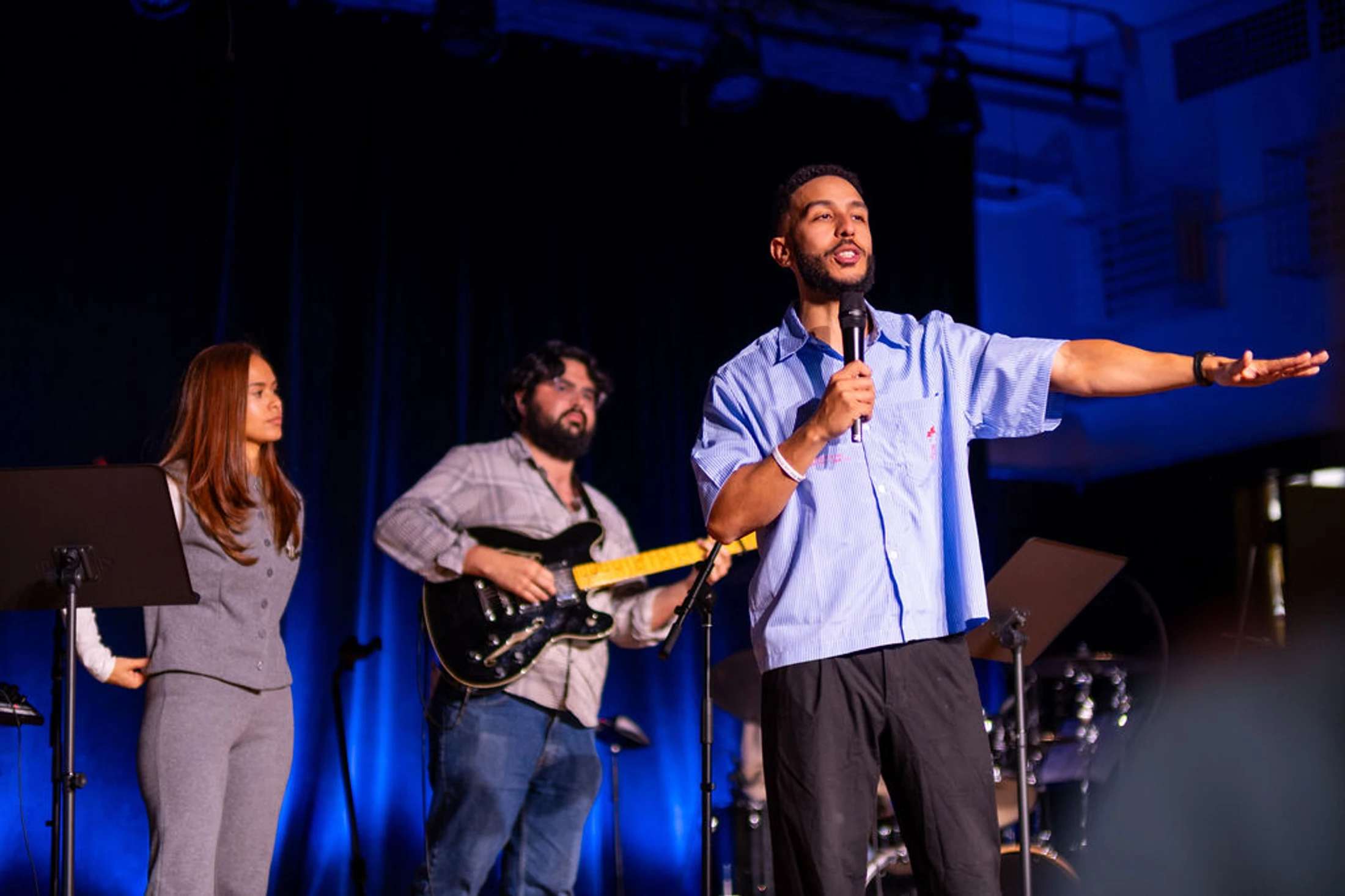 A man in a blue striped shirt speaks into a microphone and gestures to the side while on stage with a woman and a man playing an electric guitar.