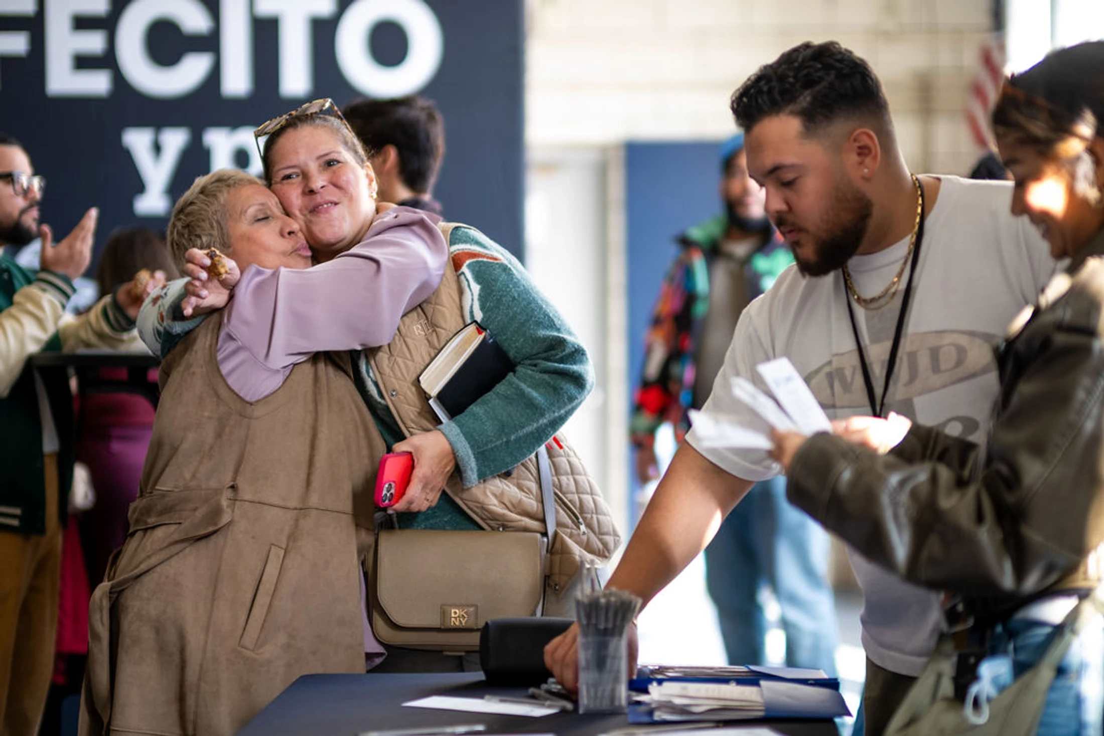 Two women embrace warmly at an event check-in table while others look at pamphlets in the foreground.