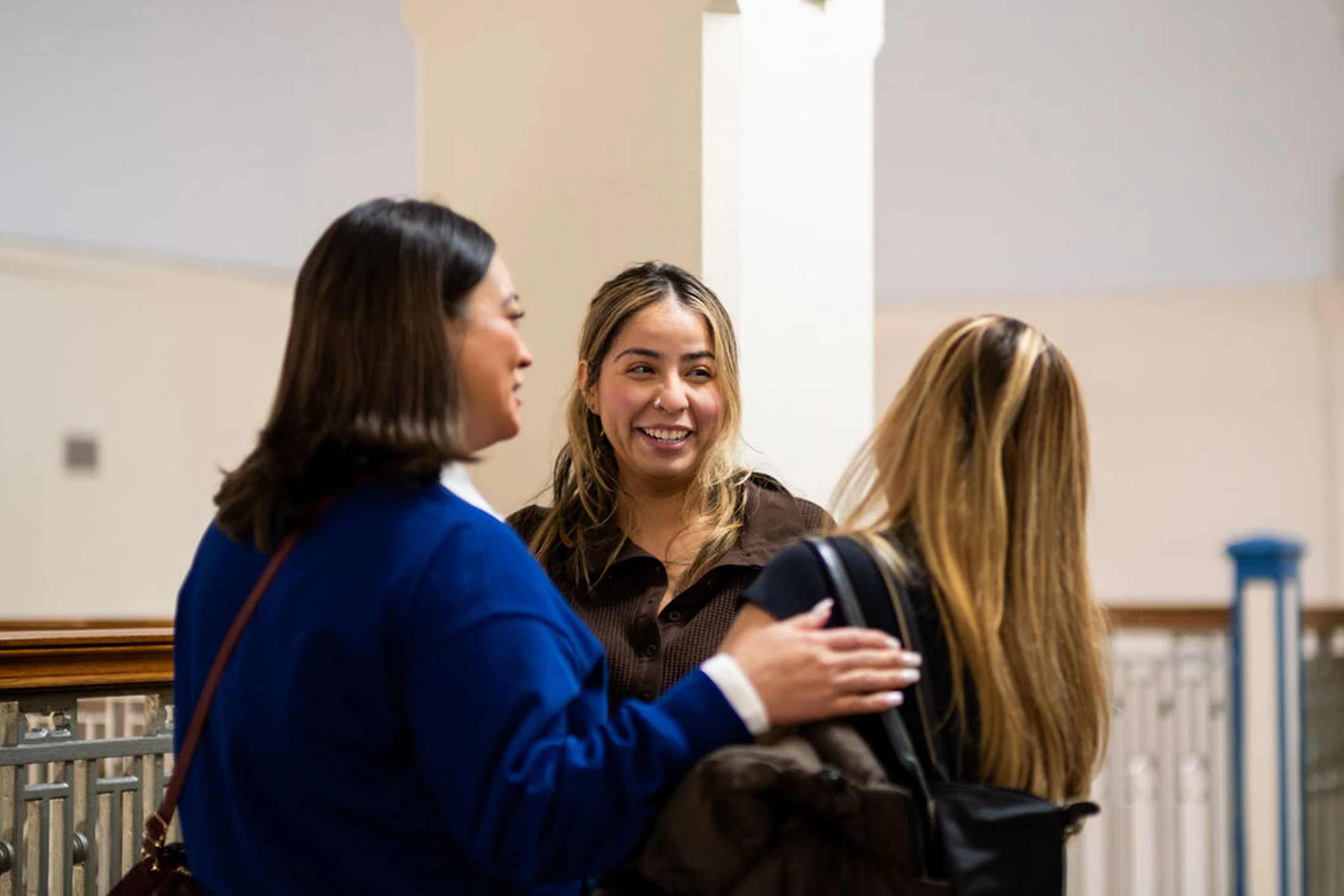 Three women stand together in a hallway. The woman in the center smiles as she talks to the woman on the left, who has her hand on the third woman's shoulder.