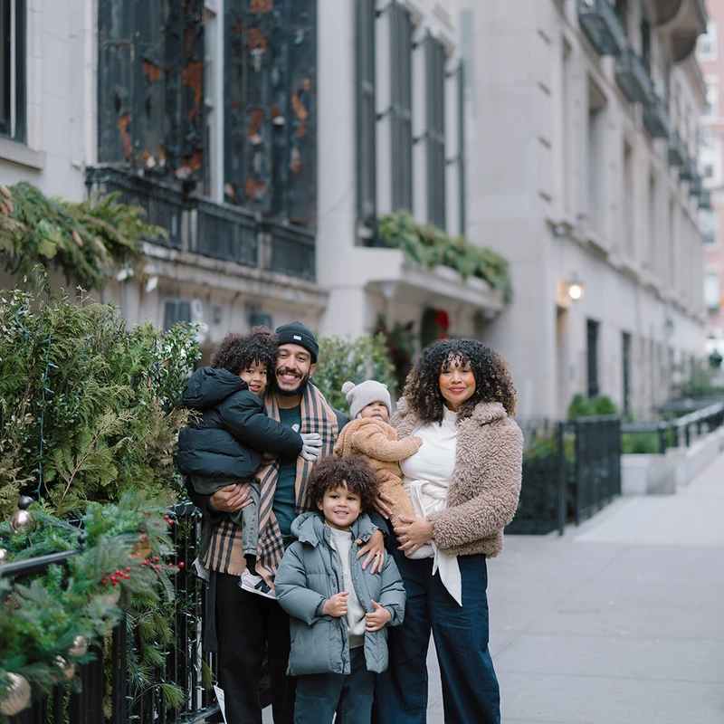 A smiling family of five, with two parents and three young sons, stand on a city sidewalk dressed in warm winter coats next to festive holiday decorations.