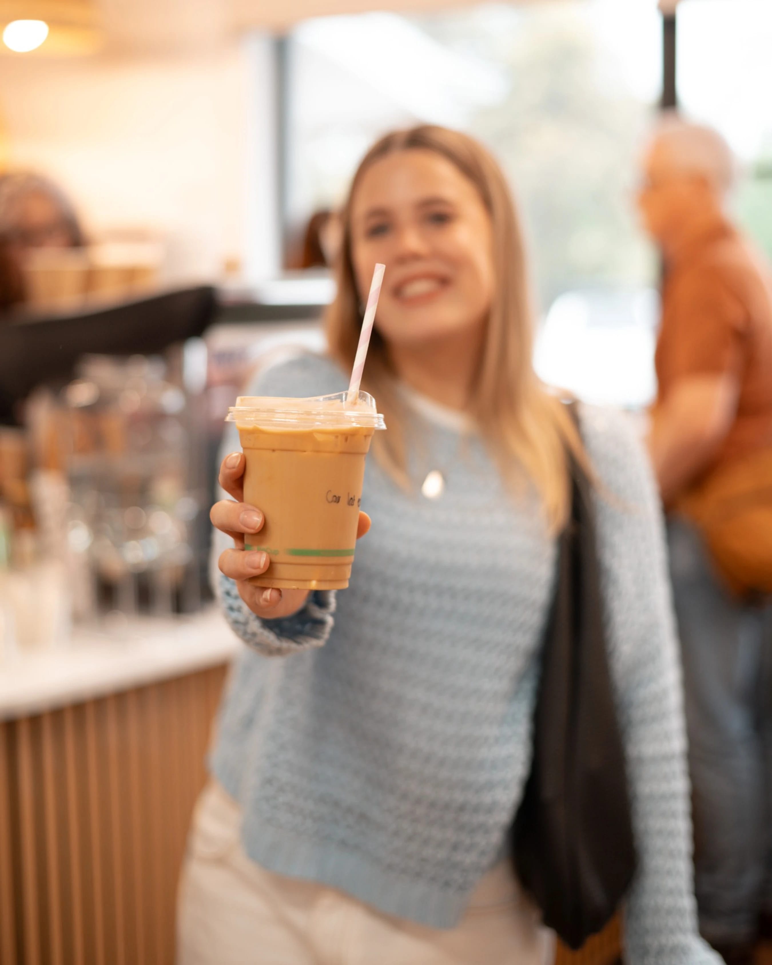 A close-up of a plastic cup of iced coffee with a pink straw, held out by a smiling woman who is blurred in the background of a coffee shop.
