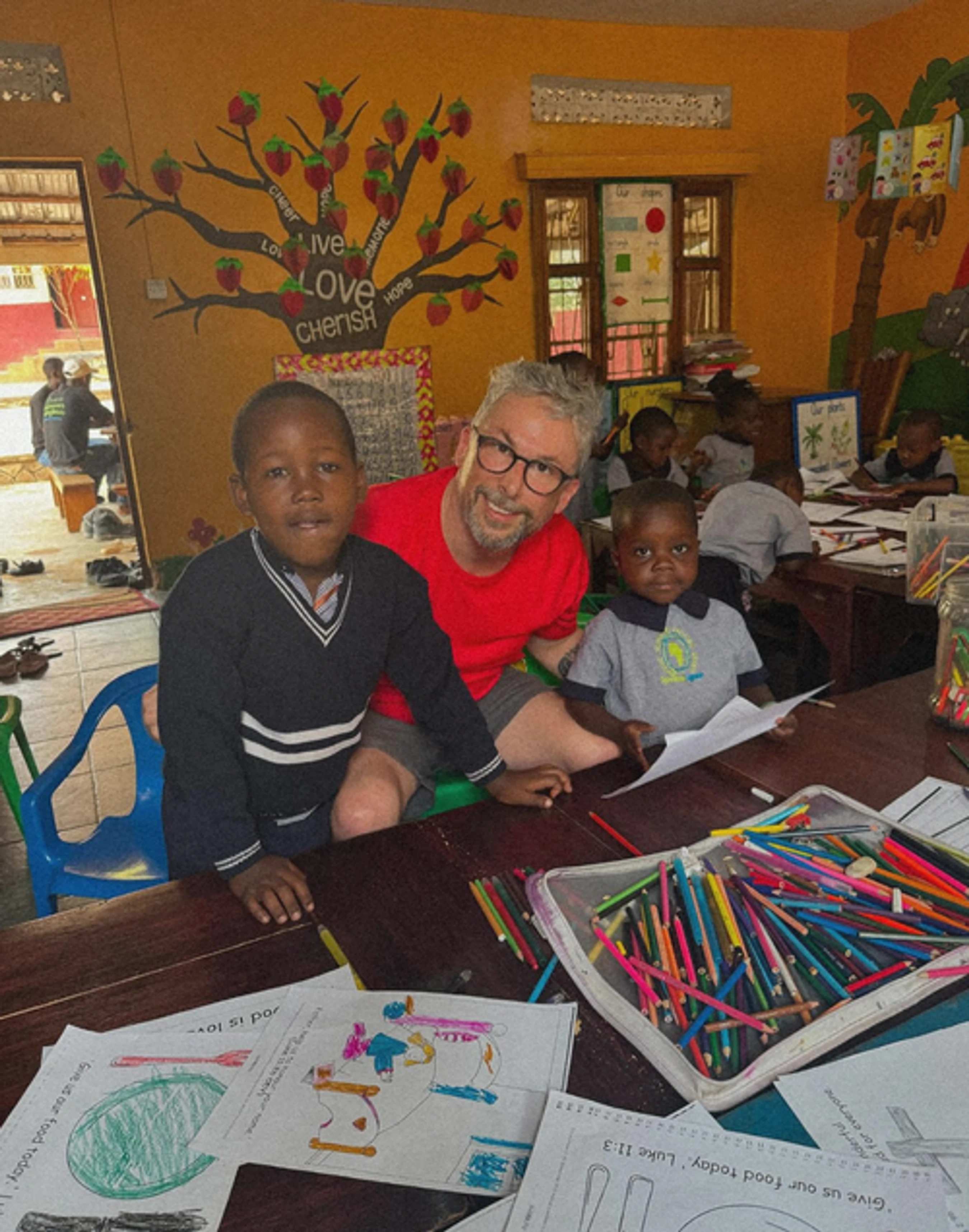 A man with glasses smiles, sitting at a table with two young boys in a colorful classroom. They have coloring sheets and many colored pencils on the table.