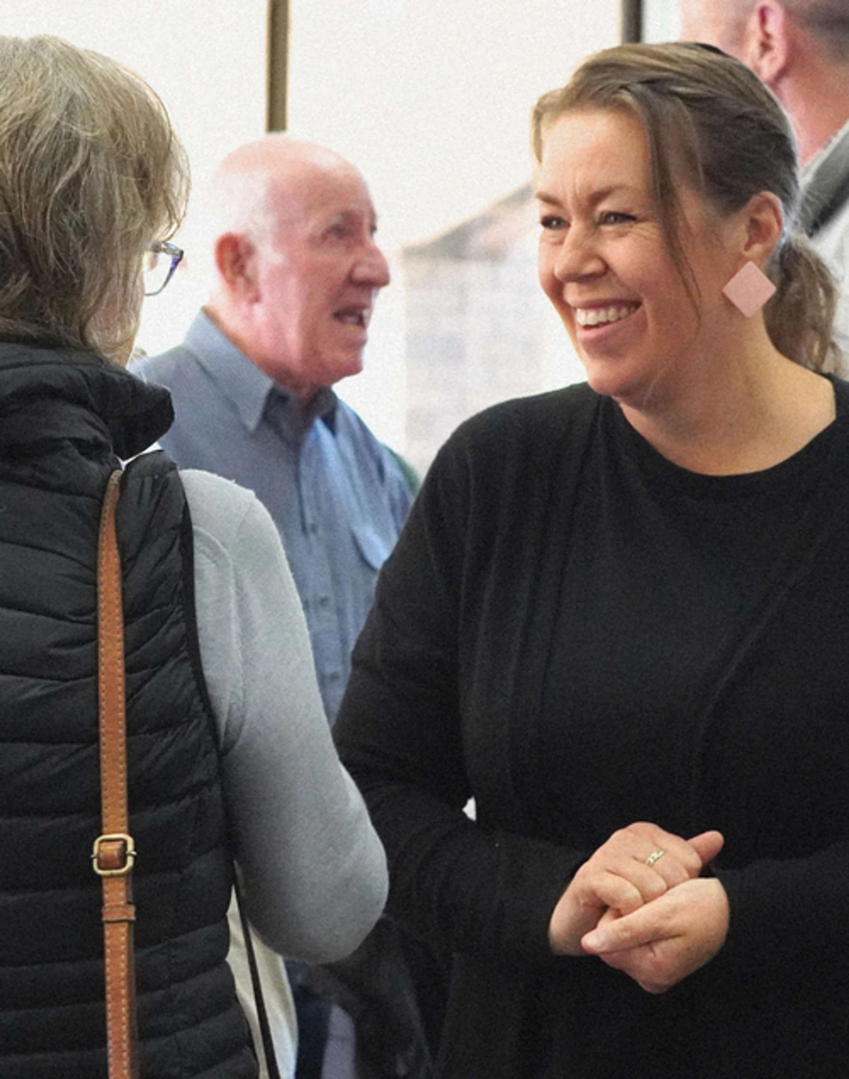 A smiling woman with her hair tied back, wearing a black top and pink earrings, engages in a friendly conversation with another person at an indoor gathering.