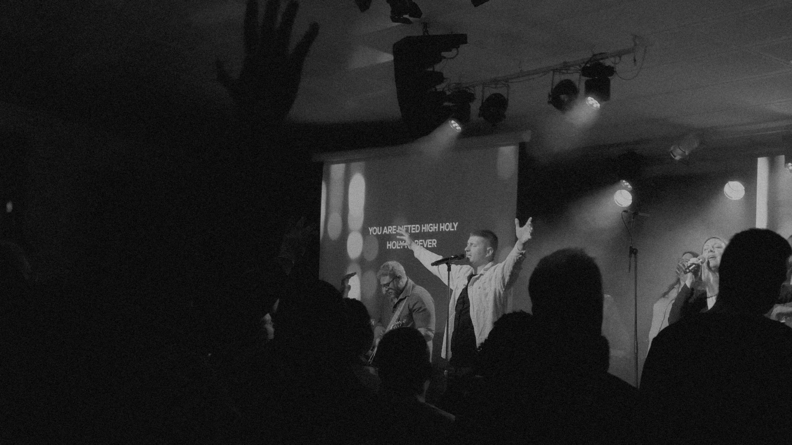 A black and white photo of a band leading worship on stage. A man sings with his hands raised as the congregation watches from the shadows.