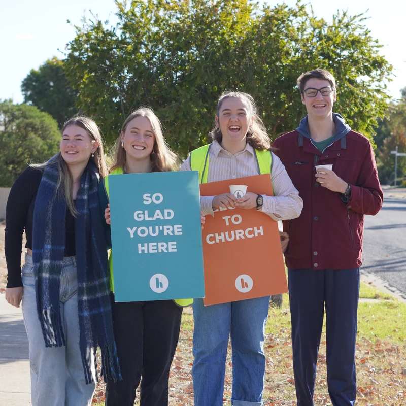 Four smiling young adults stand on a sidewalk holding welcome signs that read 'So glad you're here' and 'to Church' on a sunny day.