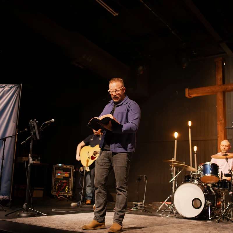 A pastor in a blue shirt and glasses stands on a dimly lit stage, reading from a Bible. A band and a large wooden cross are visible in the background.