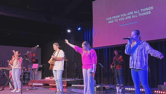 A young worship band performs on a stage with purple and blue lighting. Lyrics are displayed on a large screen behind them as they sing and play instruments.