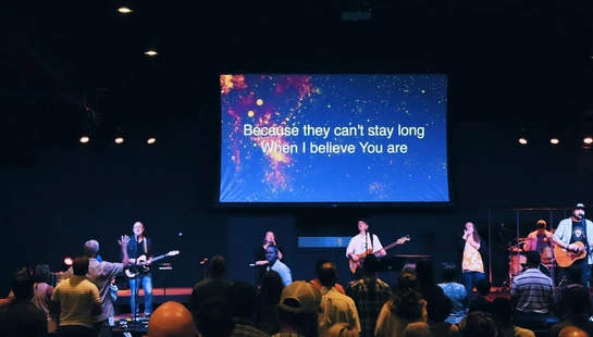 A worship band on a dimly lit stage performs for an audience. A large screen above them displays song lyrics against a blue and gold background.