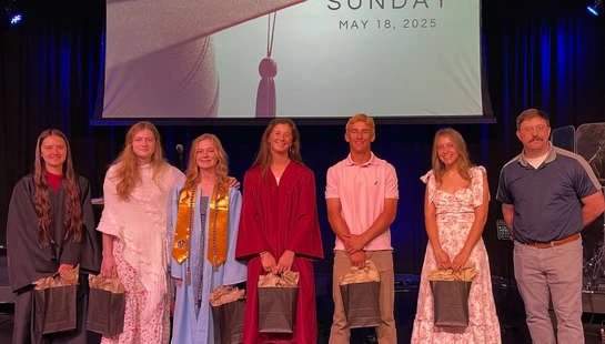 A group of six graduates and an adult stand on a stage in front of a screen that reads "Graduate Sunday." The graduates are holding gift bags.
