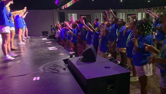 A large group of children in matching blue shirts sing and raise their hands on a stage with purple lighting, with a string of international flags above them.