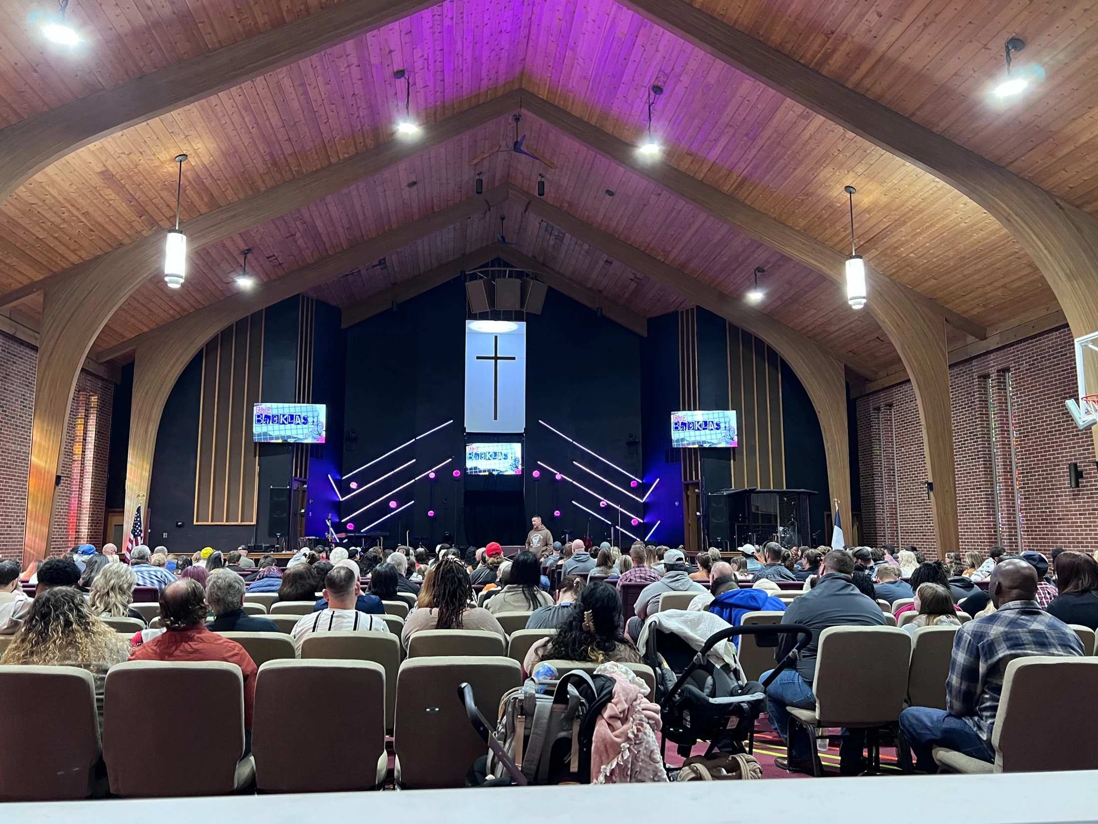 A view from the back of a crowded church, showing the congregation watching a speaker on a stage lit with purple lights, under a high, arched wooden ceiling.
