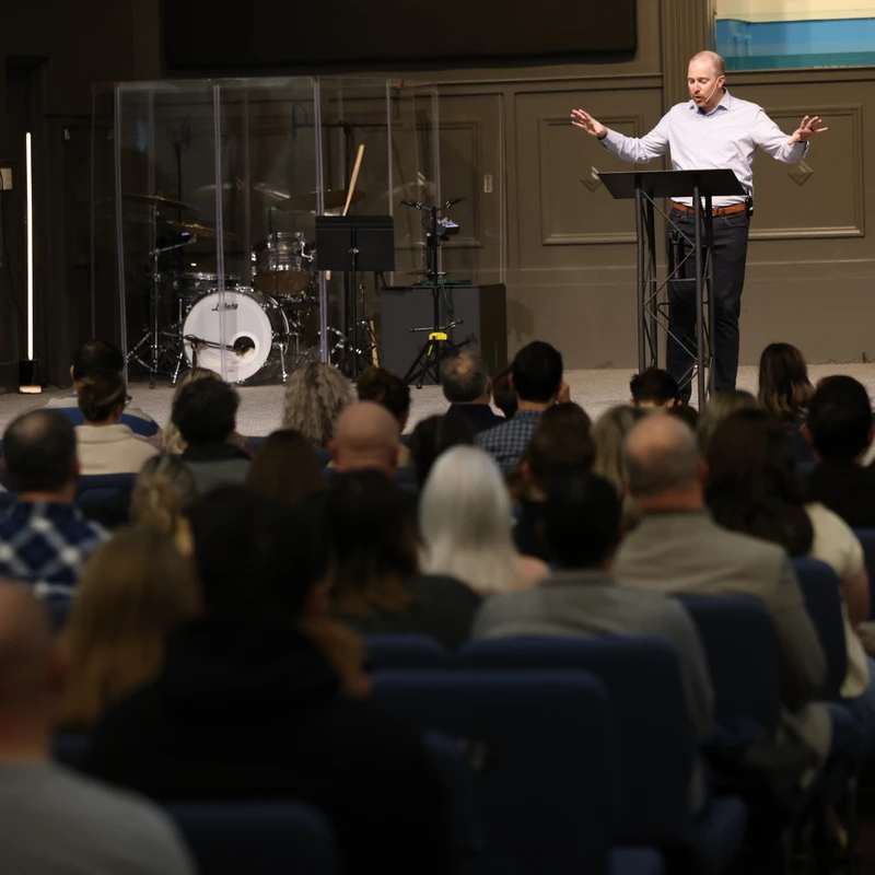 A man stands on a stage behind a podium, speaking to a large audience seated in an auditorium. A drum set is visible behind him on stage.
