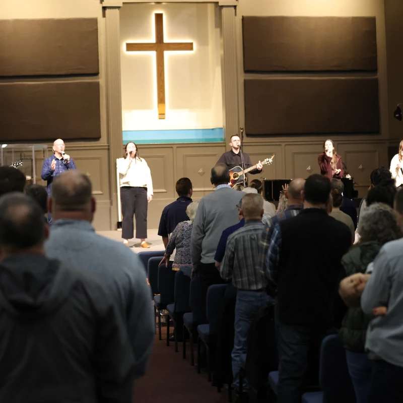 A view from the back of a church shows a congregation standing and facing a stage where a band leads worship. An illuminated cross is on the wall.