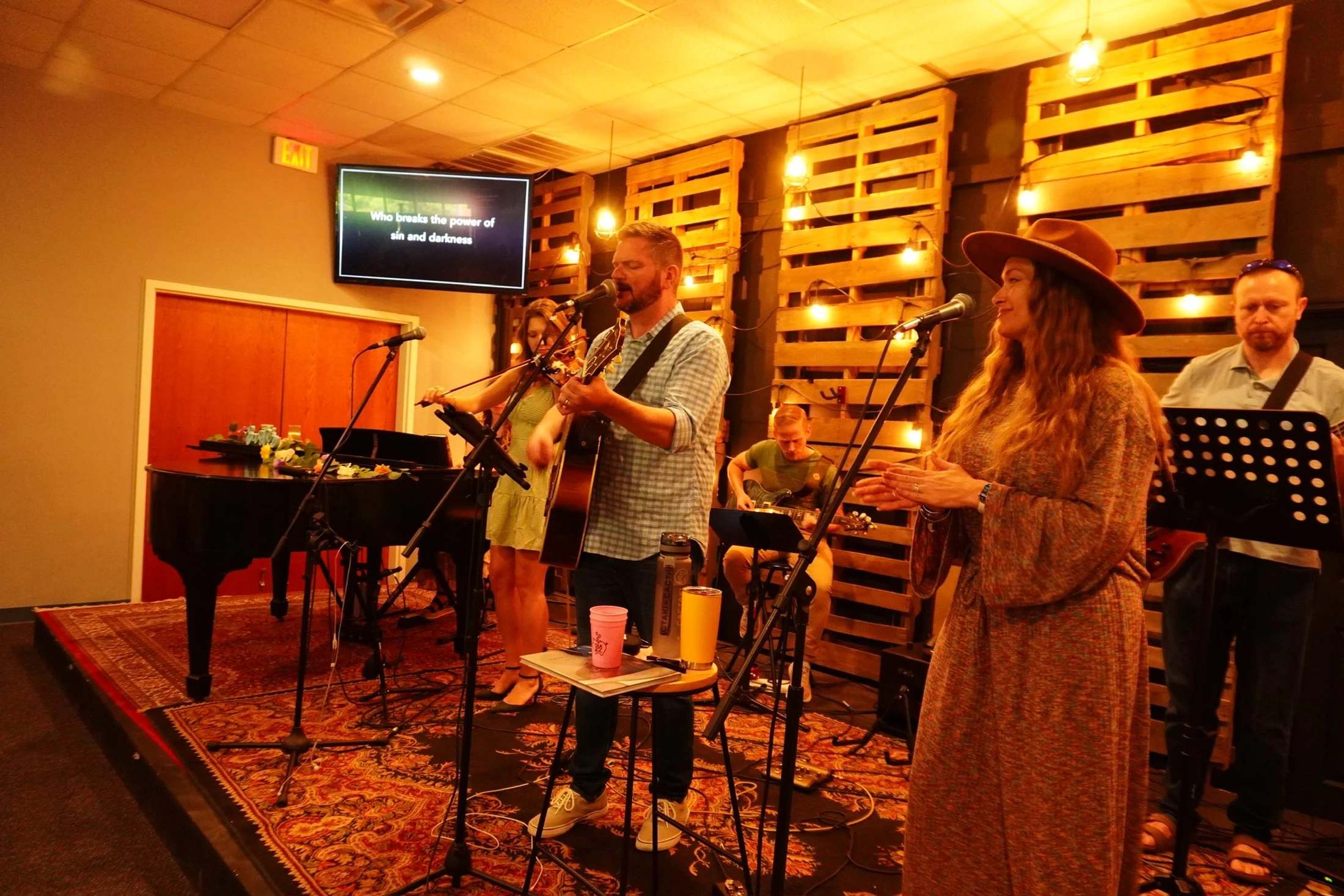 A band performs on a stage decorated with wooden pallets and warm lights. A man sings and plays guitar while a woman in a hat sings into a microphone.