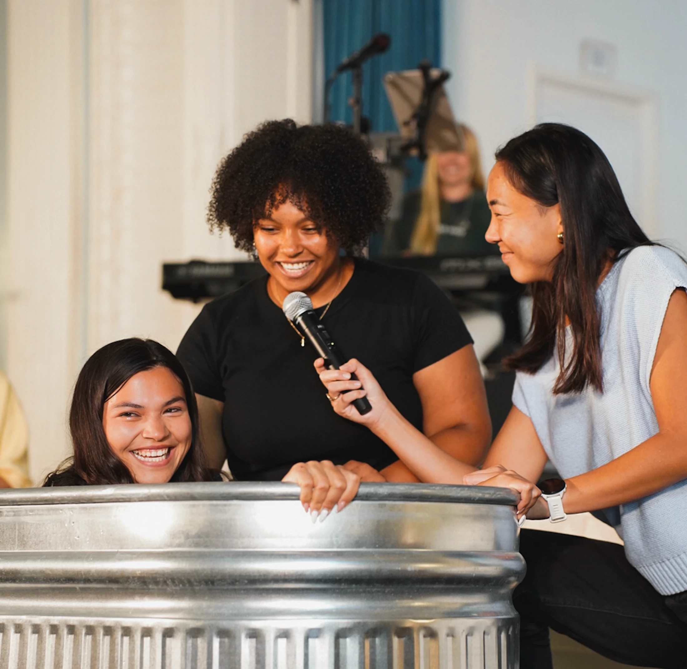 A smiling woman sits in a large metal tub for her baptism while two other women stand beside her, one holding a microphone for the other to speak into.