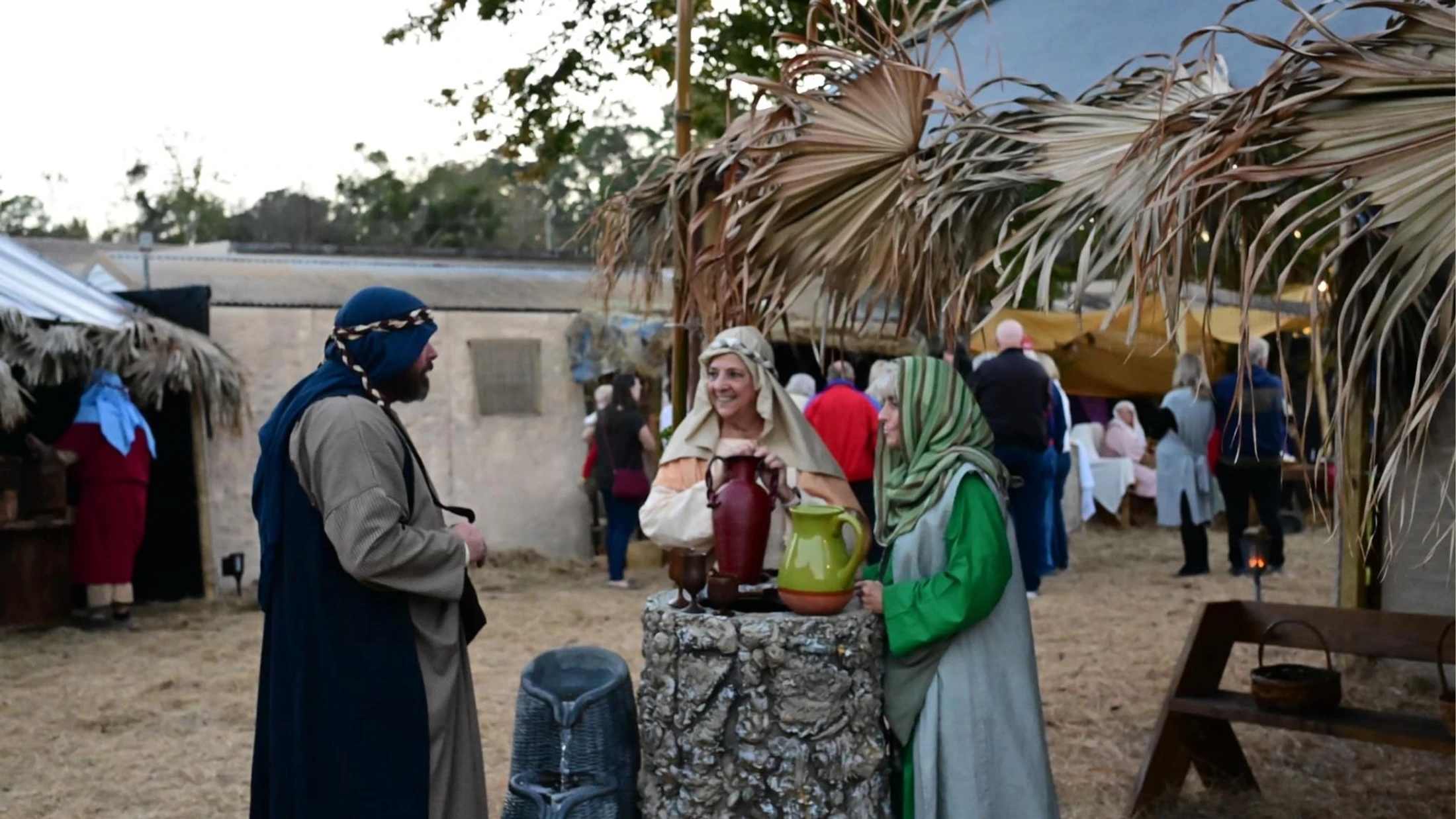 Three people in biblical-era costumes stand around a stone well with pottery jugs, participating in an outdoor historical or religious reenactment.
