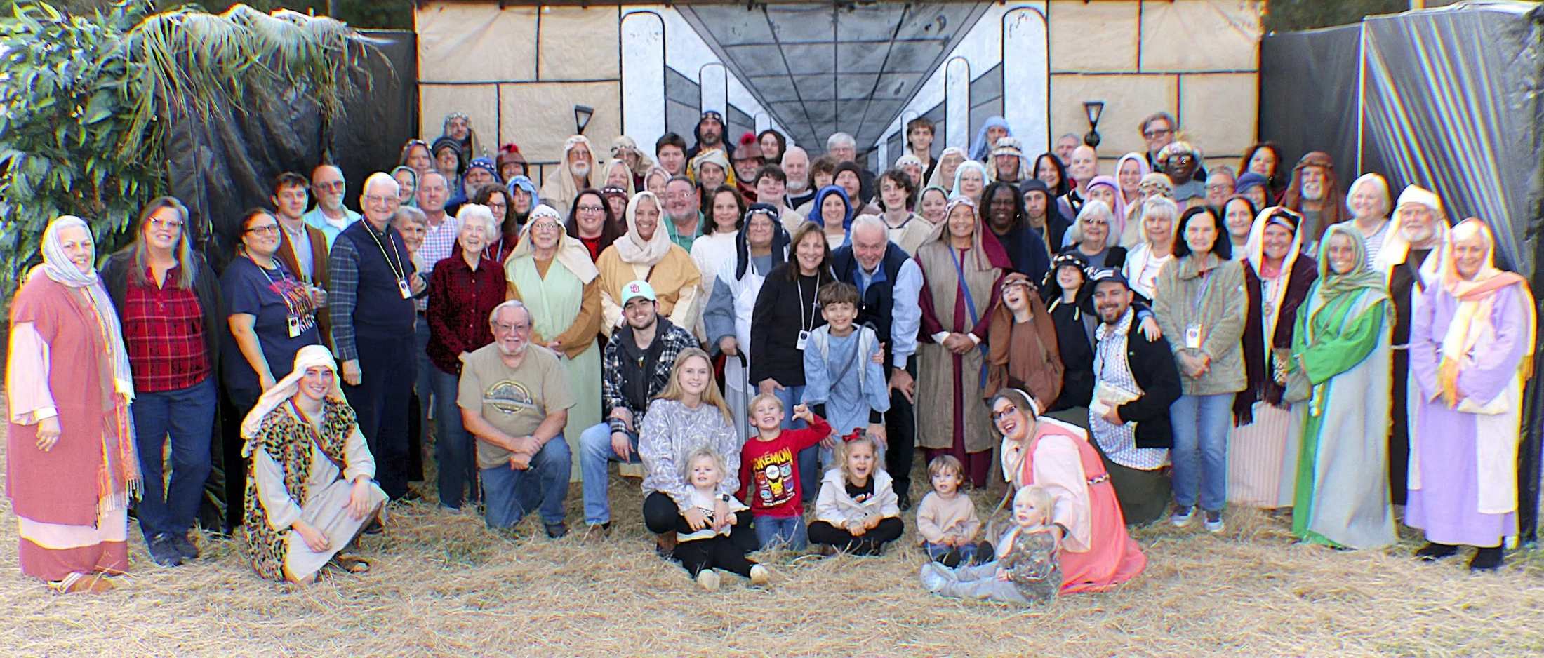 A large, multi-generational group of people, many in biblical-style costumes, pose for a photo outdoors on hay in front of a stage set.
