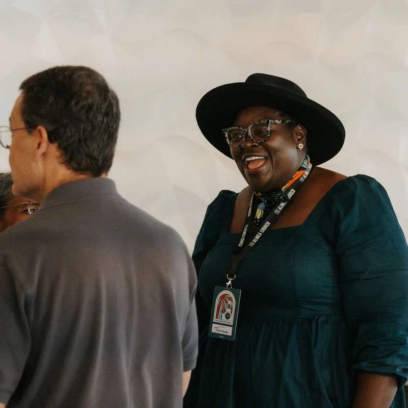 A Black woman wearing a black hat and glasses smiles happily while talking with someone. She is wearing a volunteer lanyard and a dark blue dress.